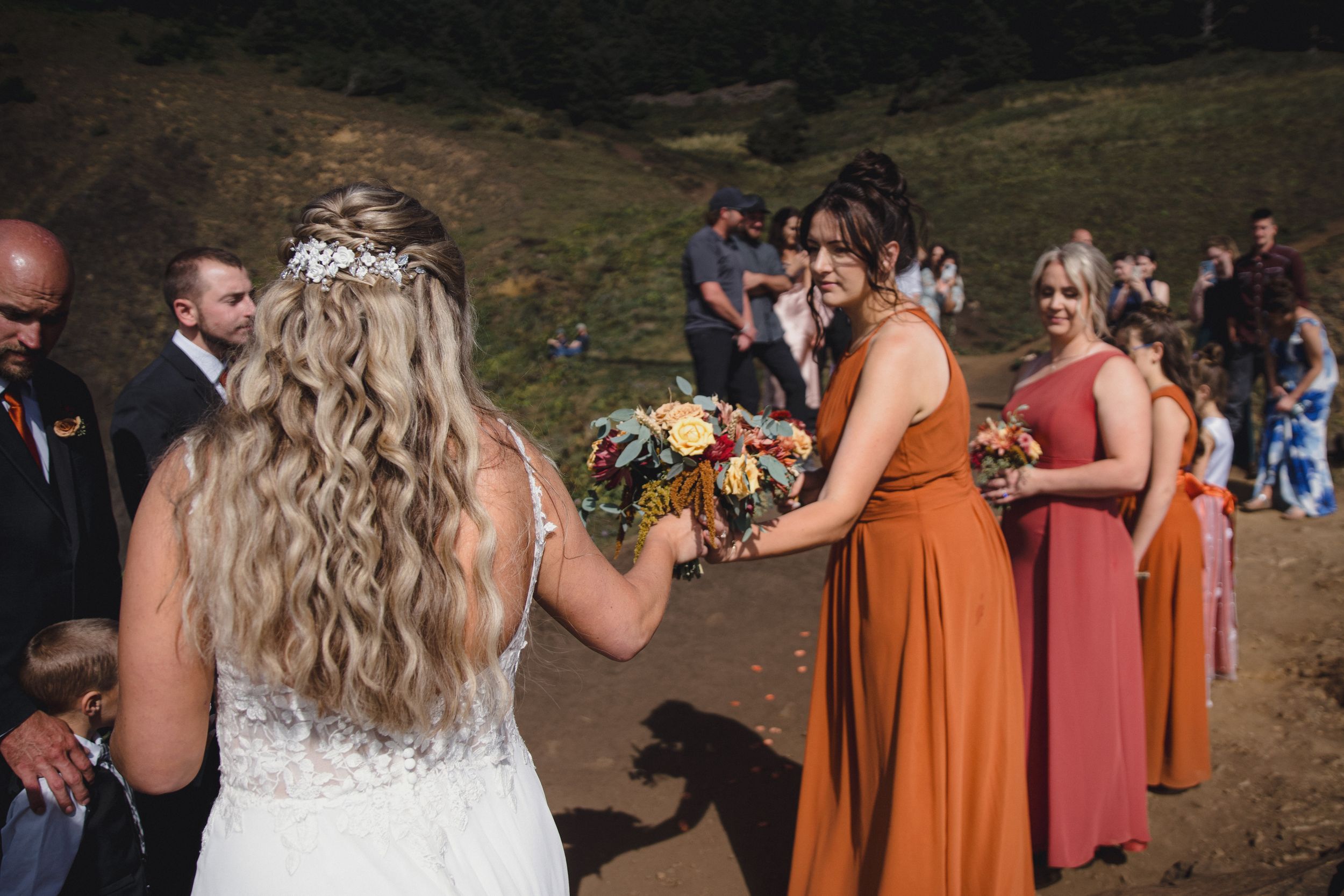 Bride in white gown receives bouquet from bridesmaid in rust orange dress at outdoor wedding ceremony.
