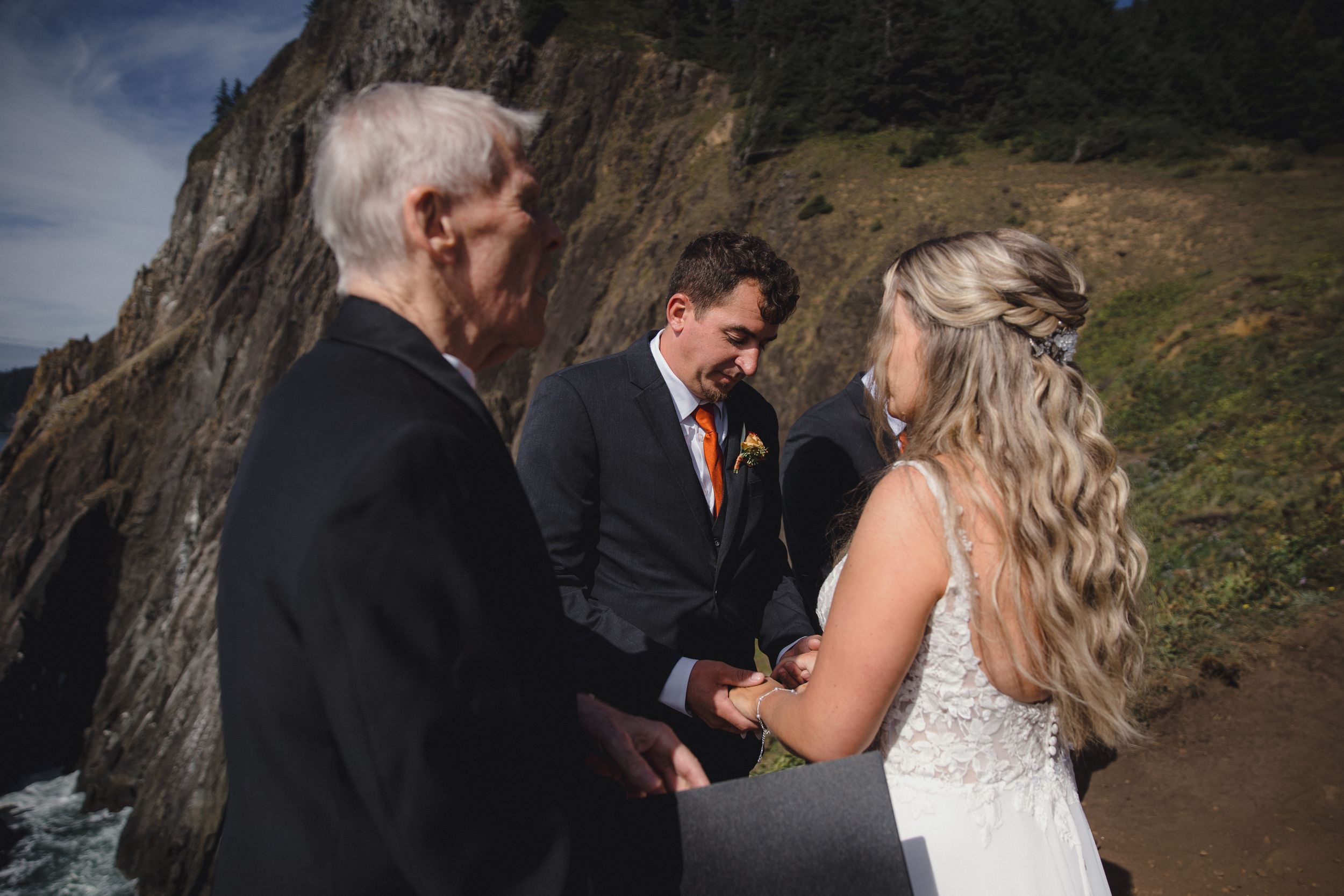 Couple exchanging vows at outdoor coastal wedding ceremony with officiant, dramatic cliffside ocean backdrop.