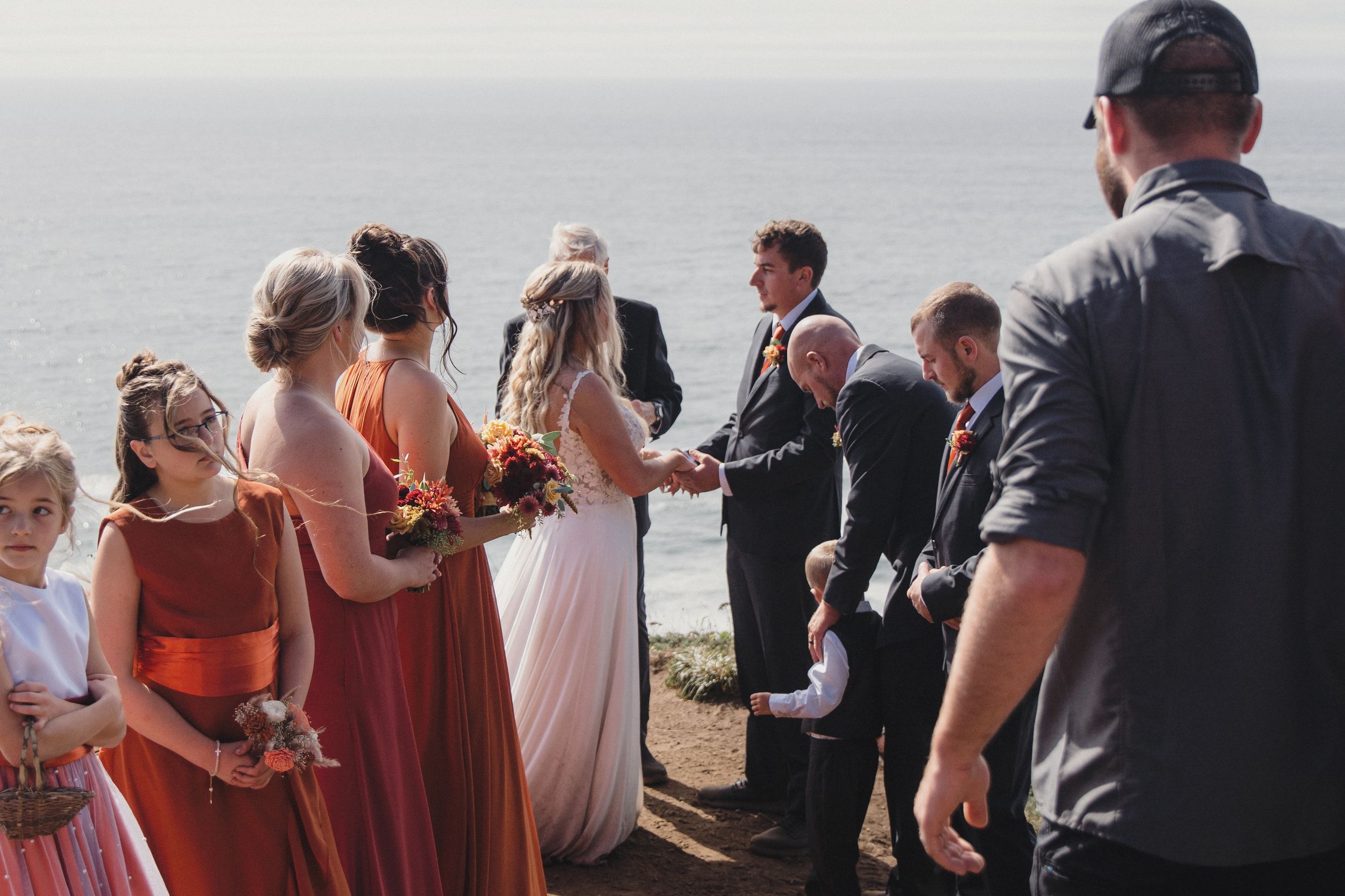 Outdoor cliffside wedding ceremony with bridesmaids in coral dresses, bride in white, and ocean backdrop.