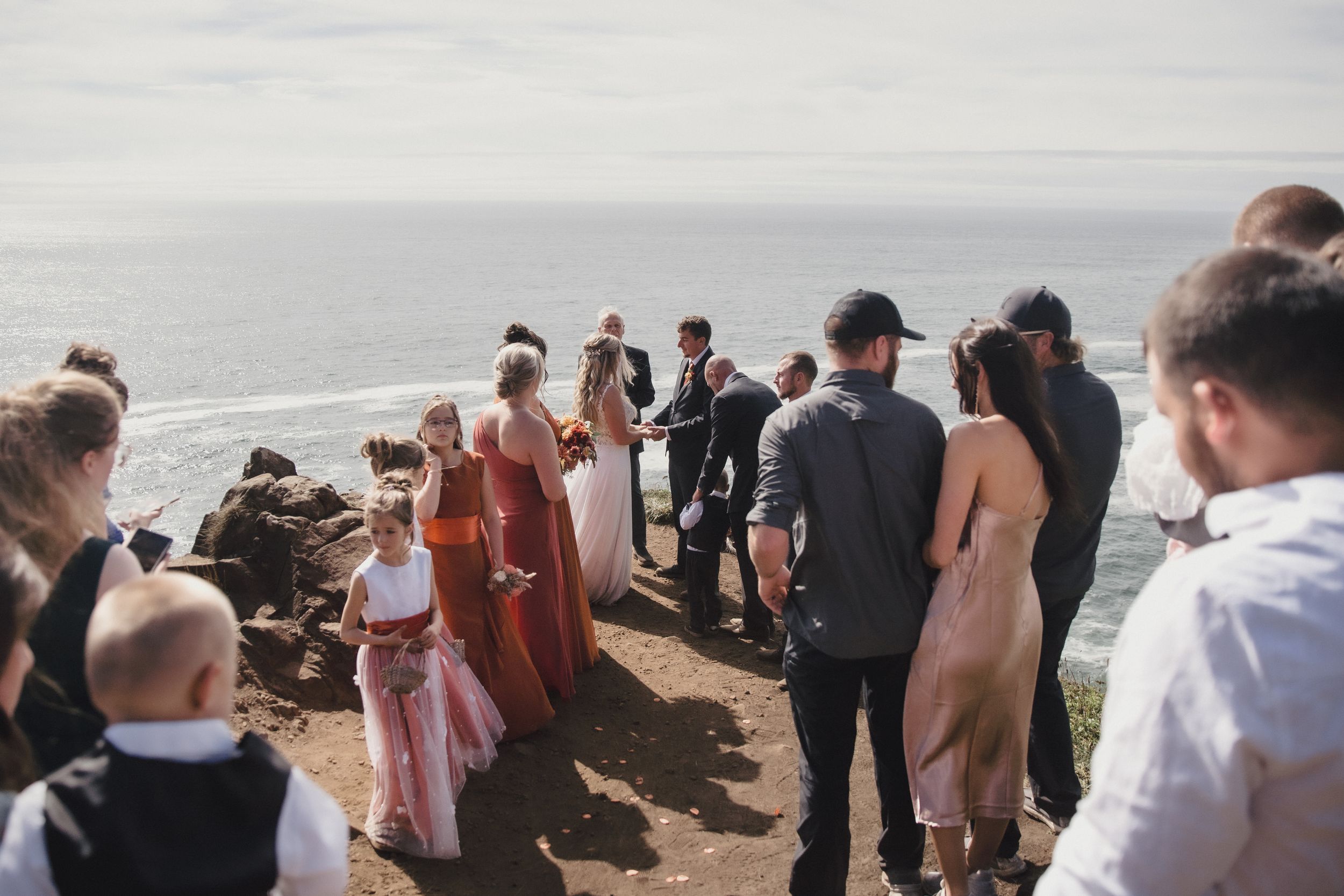Wedding ceremony on a scenic coastal cliff with guests in formal attire, ocean backdrop, sunny day.