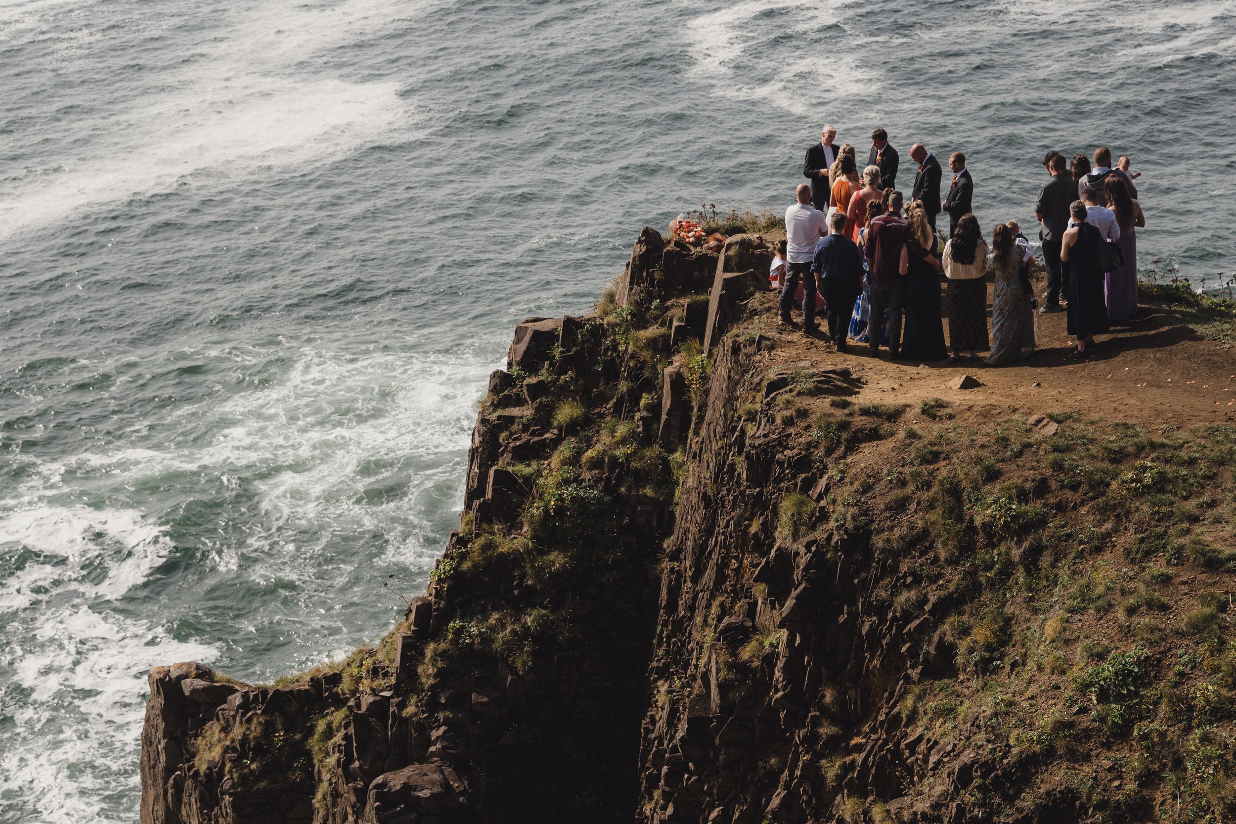 Group of tourists standing on a rocky clifftop overlooking dramatic ocean waves below.