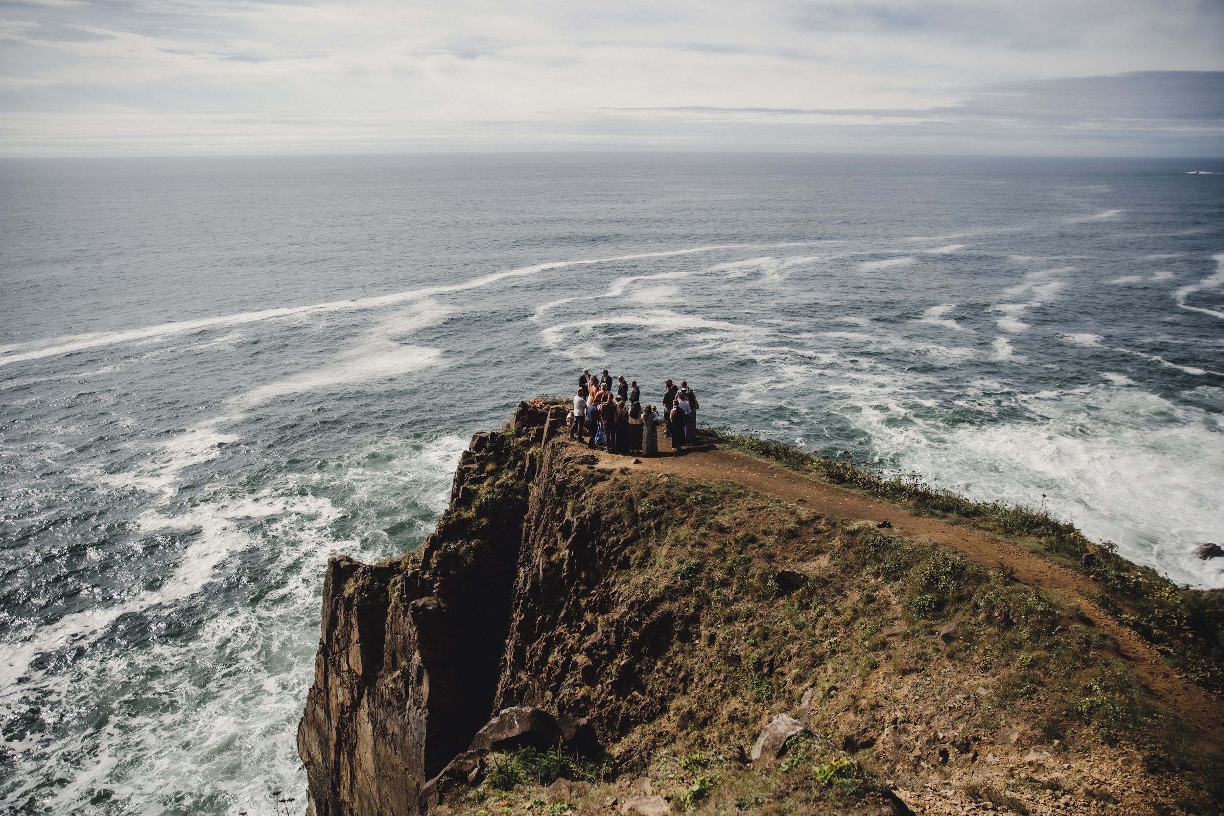 Group of people standing on a dramatic coastal cliff overlooking crashing ocean waves on a cloudy day.