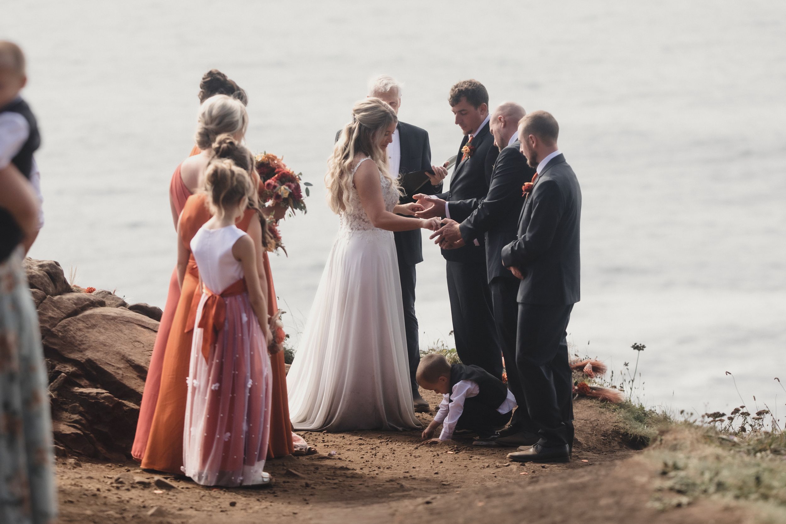 Outdoor cliff wedding ceremony with bride and groom exchanging vows, bridesmaids in pink dresses, ocean backdrop.