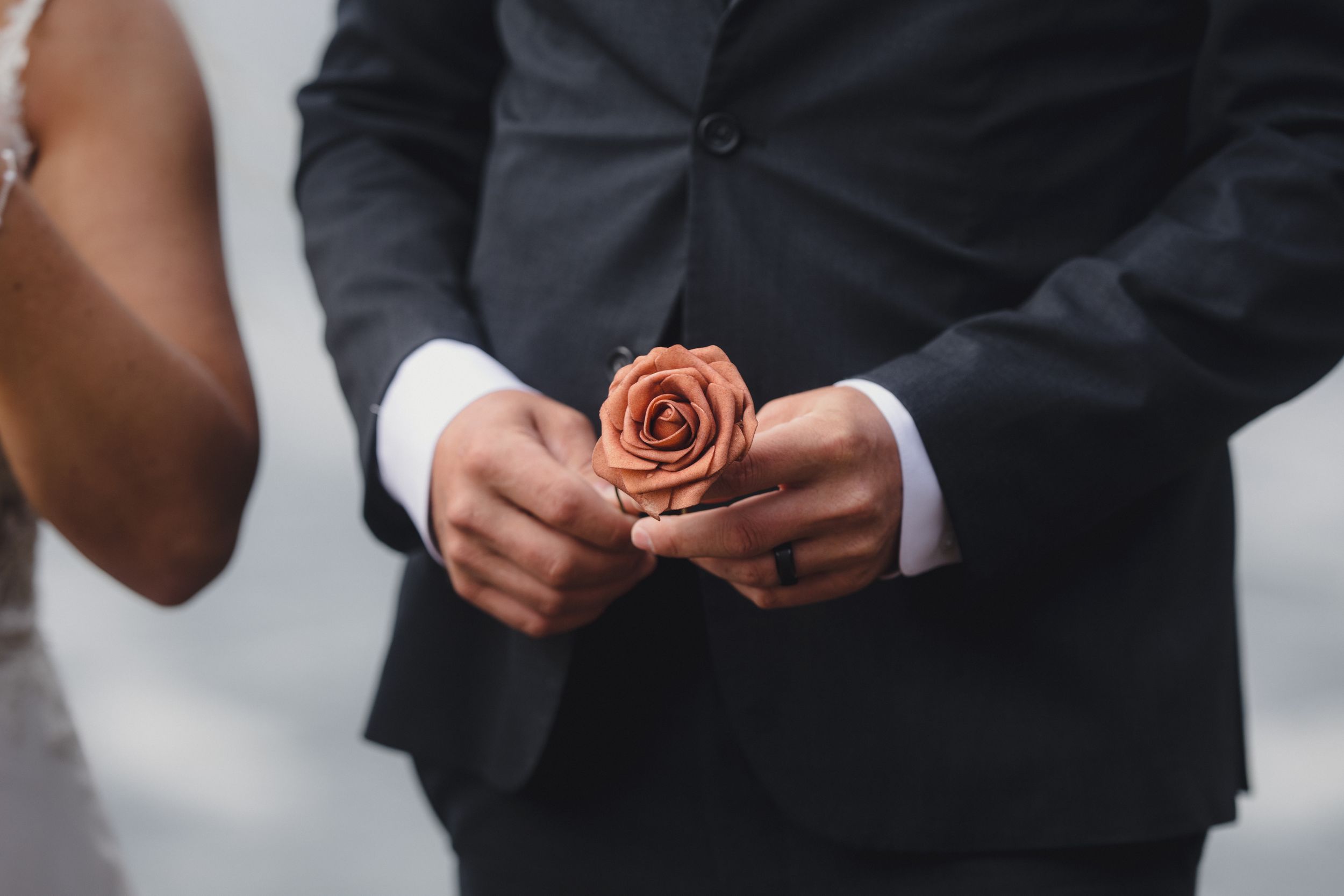 A groom in a black suit holds a terracotta rose boutonniere at a wedding ceremony.