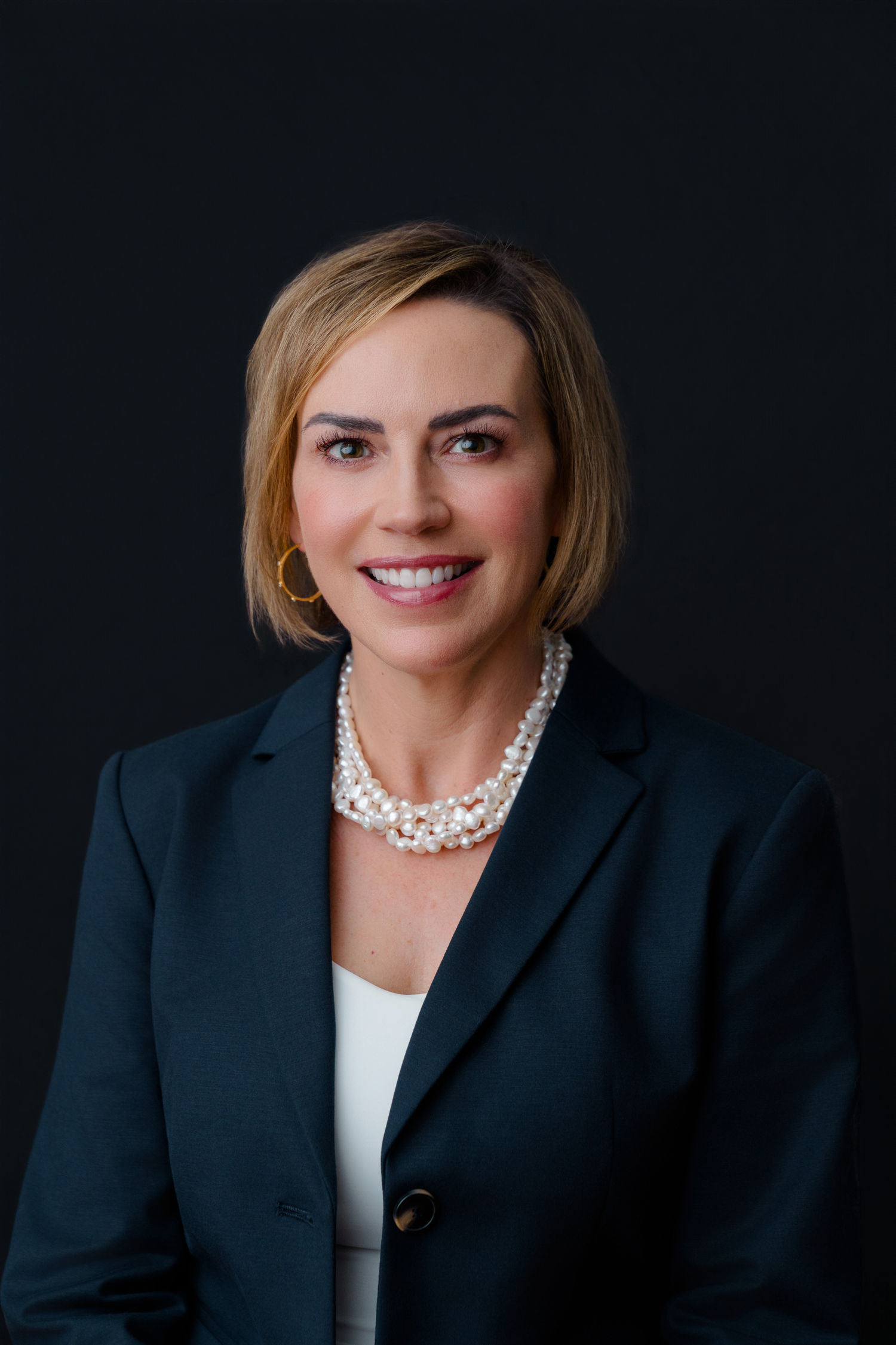 Professional woman in dark blazer and pearl necklace smiling confidently against dark background.