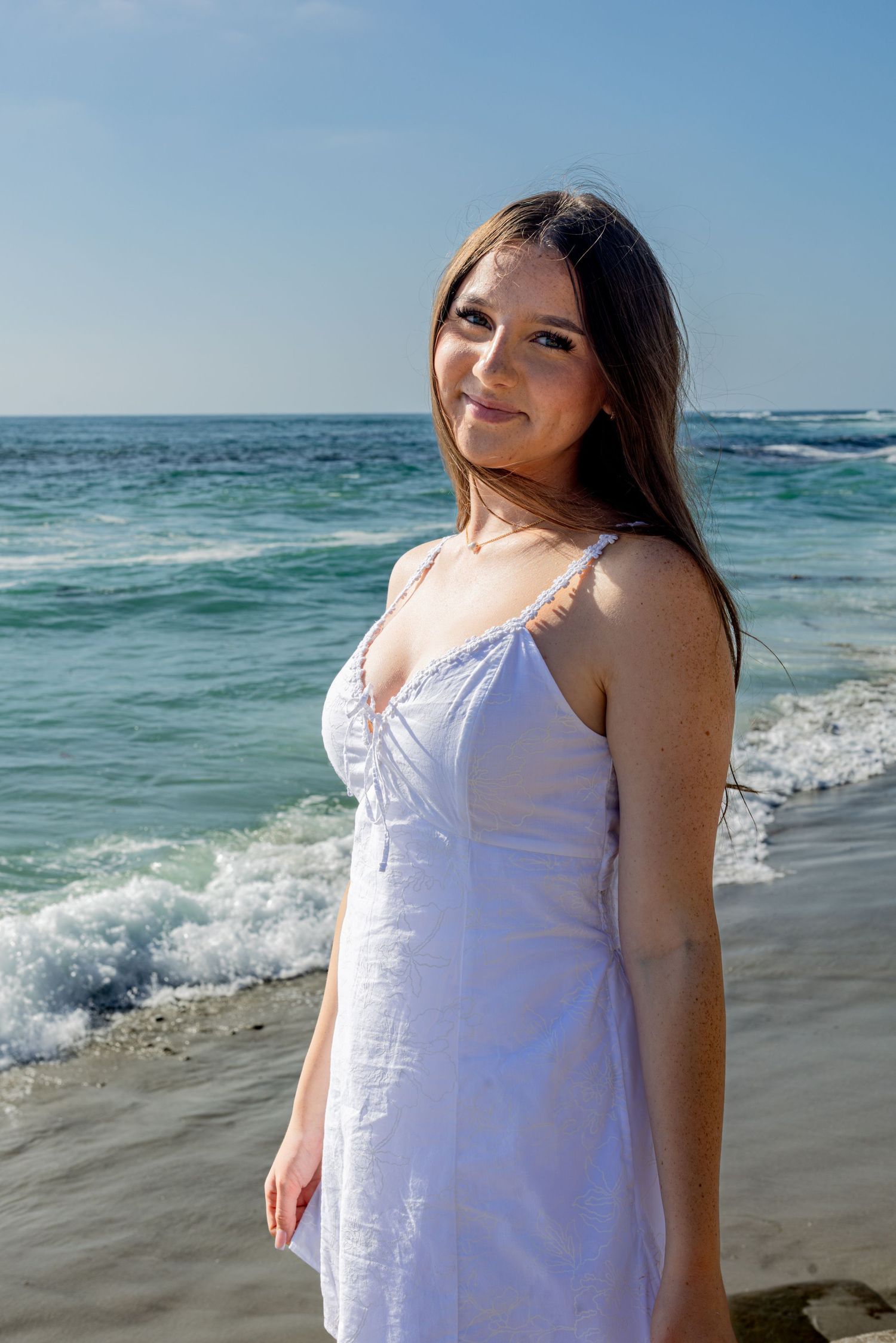 San Diego beach portrait photography of a young woman in a white sundress posing in front of crashing ocean waves at Windansea Beach.