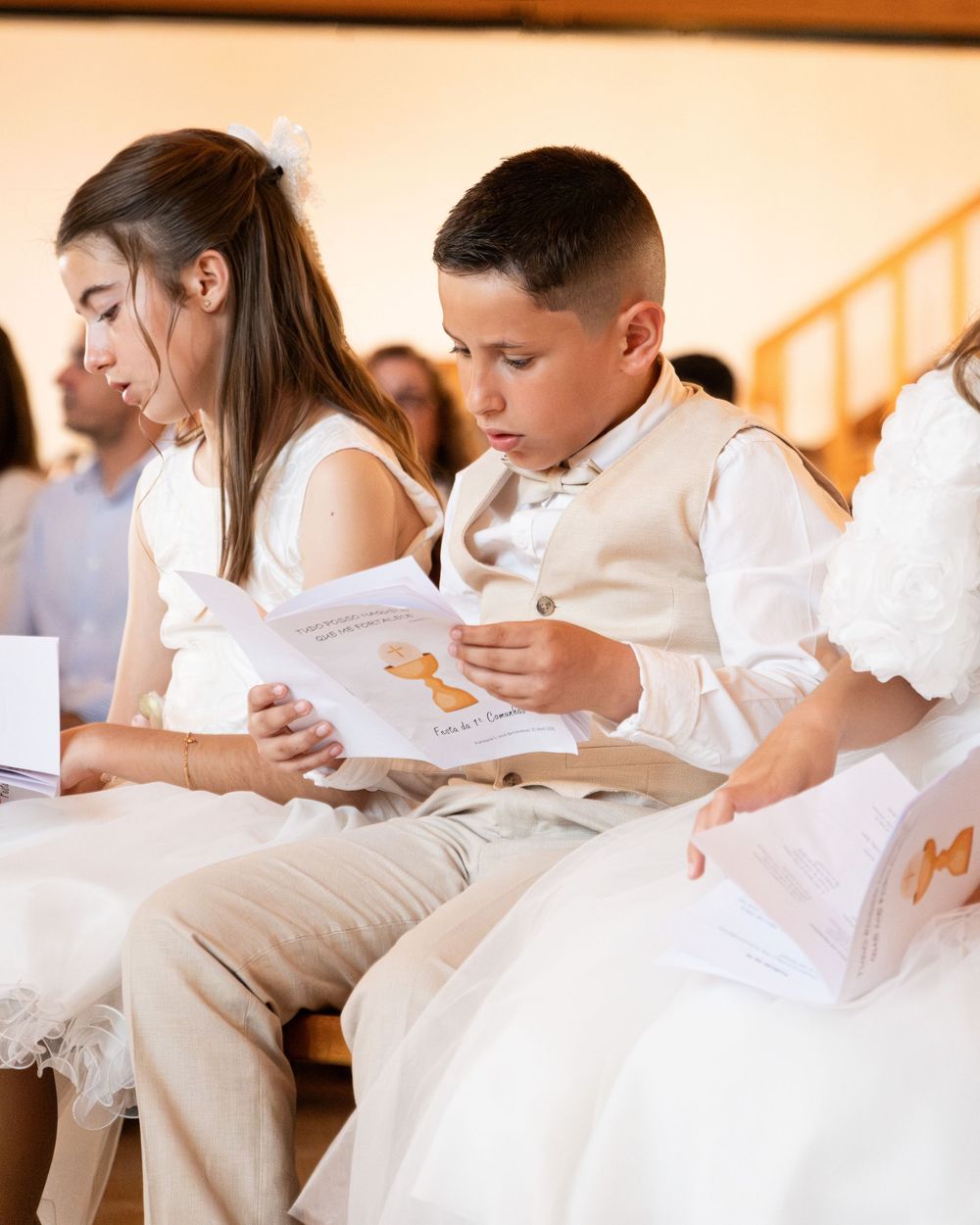 Children in white formal attire reading booklets during a First Communion ceremony in a church.