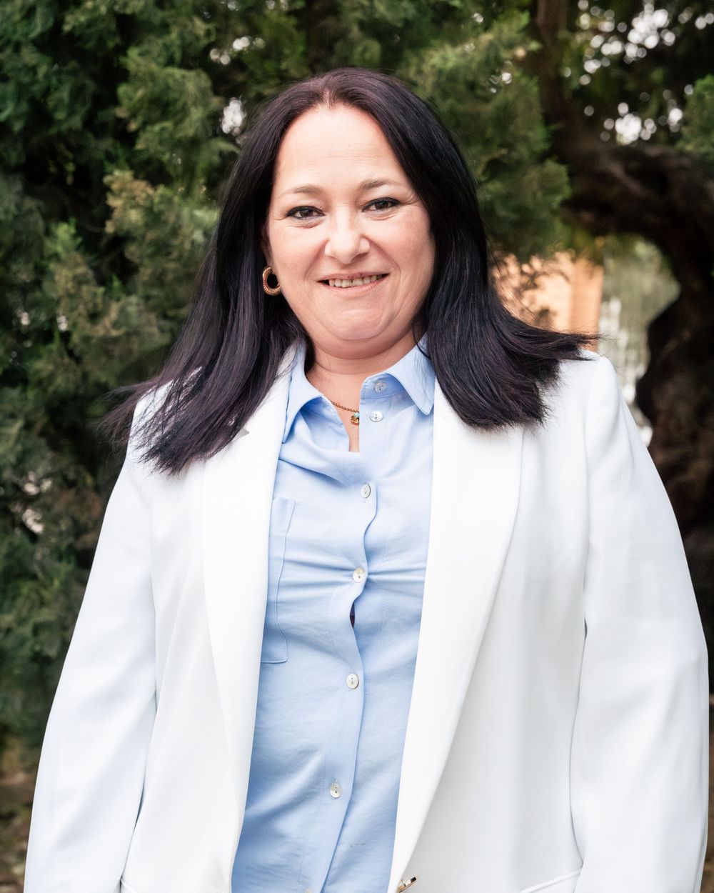 Professional woman in white blazer and blue shirt smiling outdoors with green trees in background.