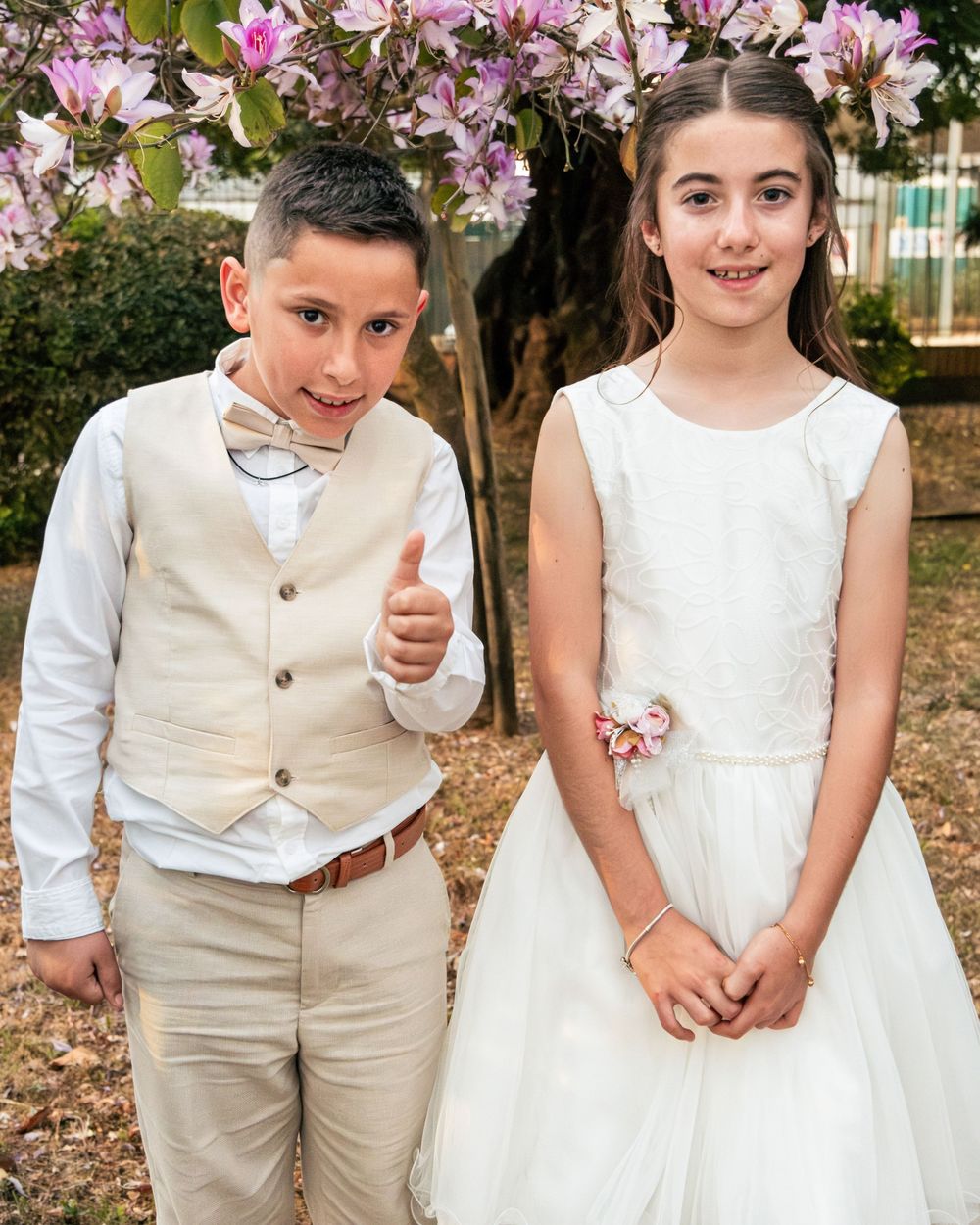 Two children dressed in formal wedding attire pose outdoors under blooming pink flowers, boy giving thumbs up.