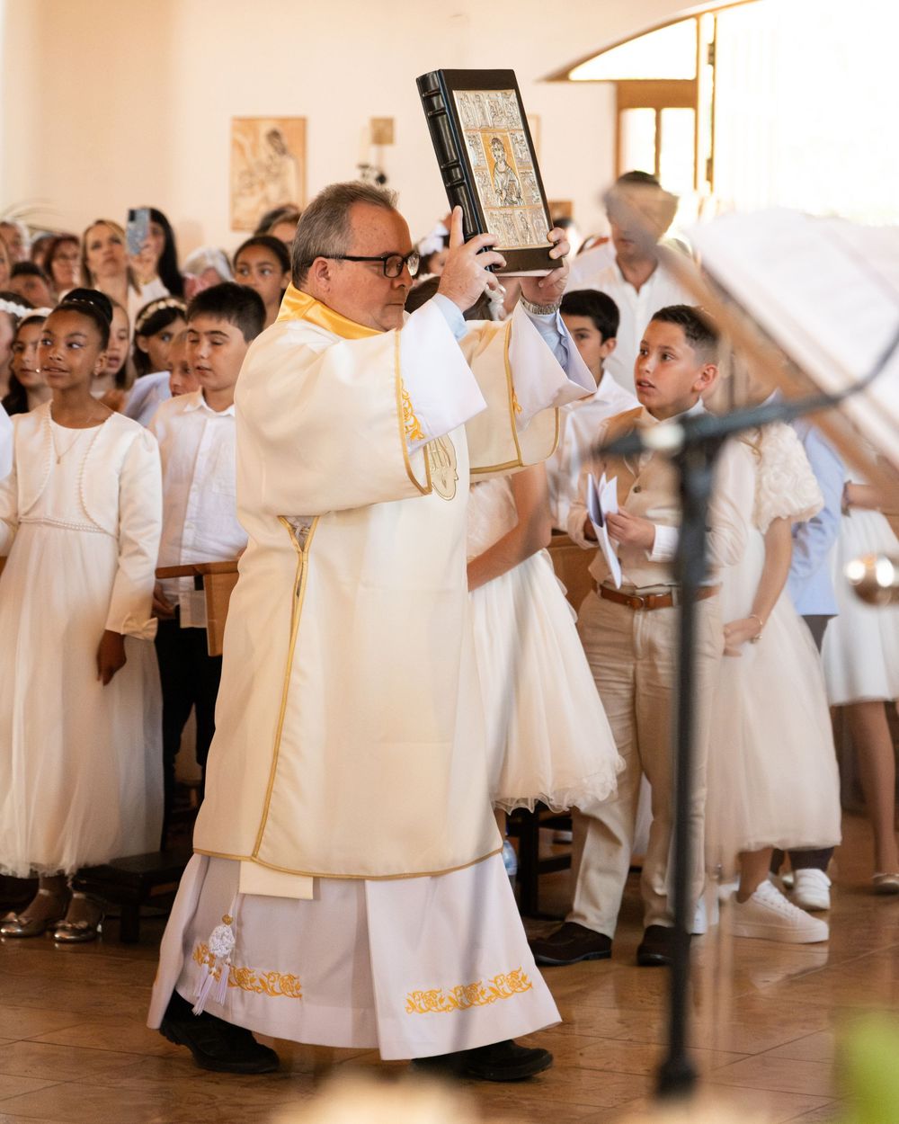 Priest in white vestments raises a holy book during First Communion ceremony with children in white attire in church.