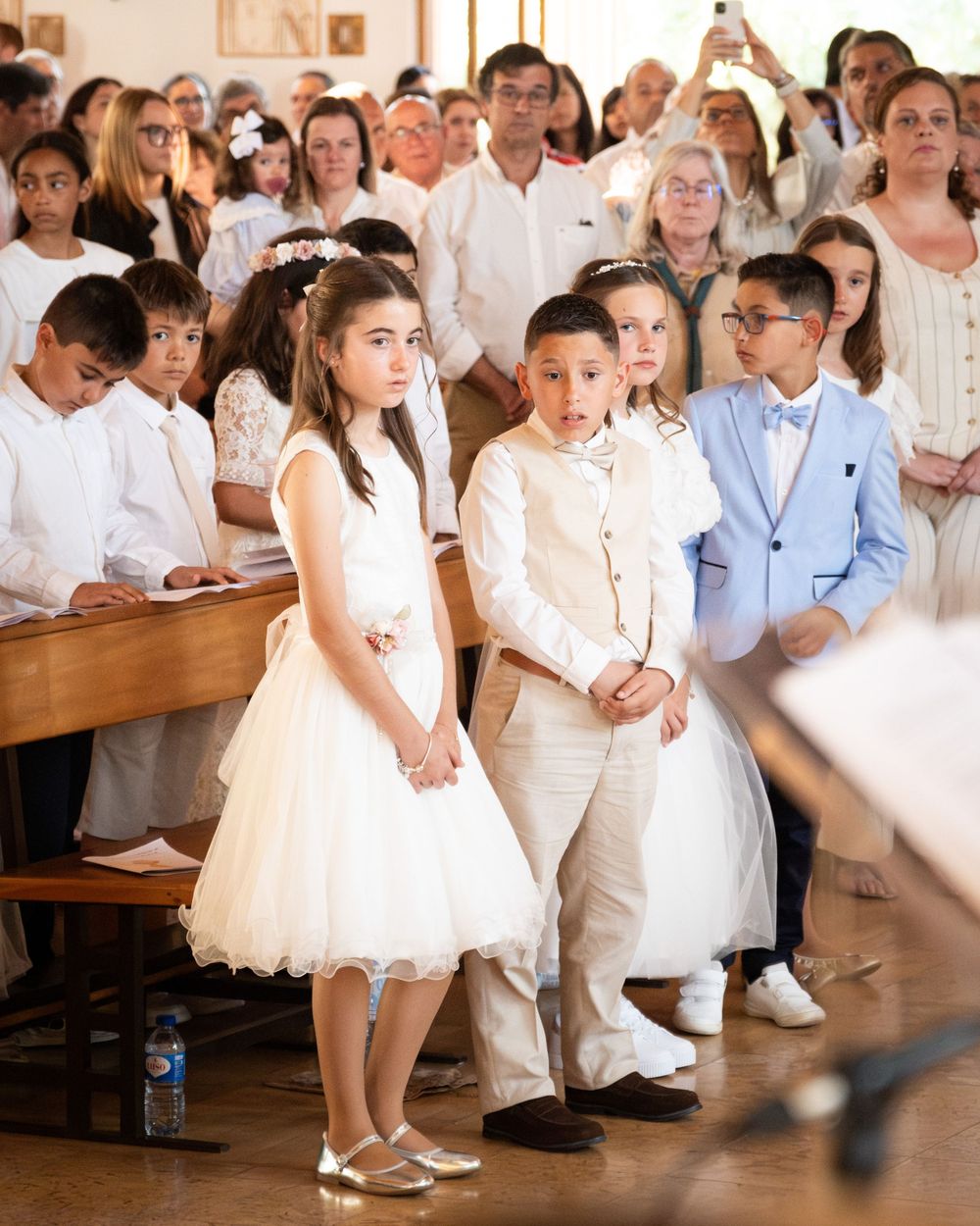 Children in formal white attire stand during a First Communion ceremony inside a church with adults seated behind.