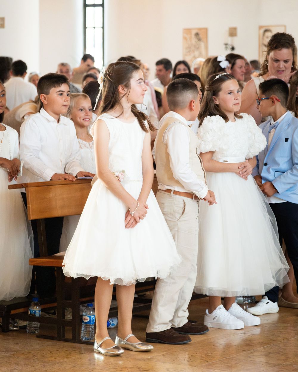 Children in white formal attire stand together during a First Communion ceremony inside a church.