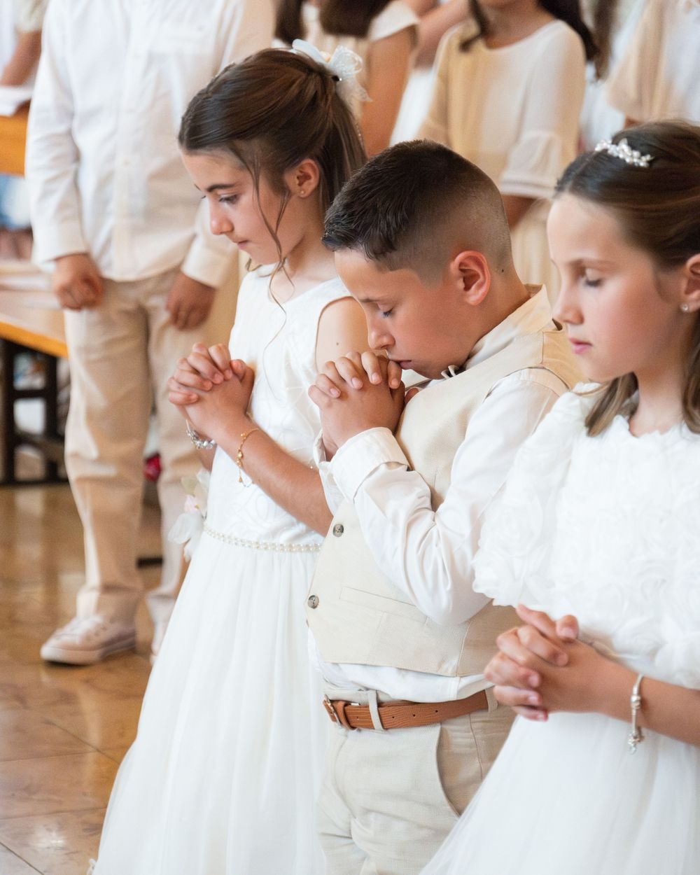 Children dressed in white pray with clasped hands during a First Communion ceremony in a church.