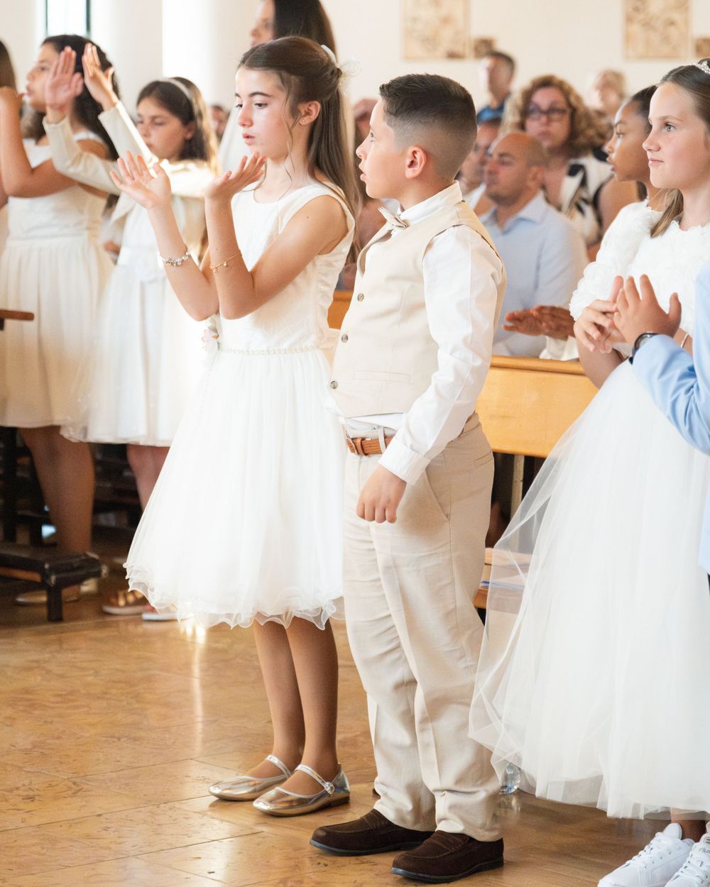 Children dressed in white formal attire participate in a First Communion ceremony inside a church.