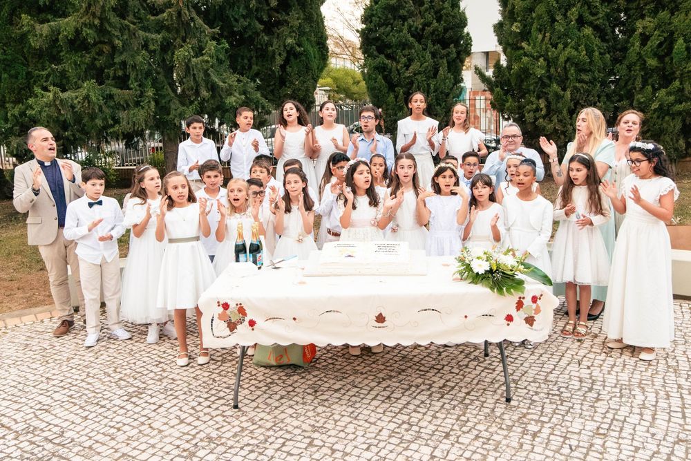 Group of children and adults in white attire posing together at a First Communion celebration outdoors.