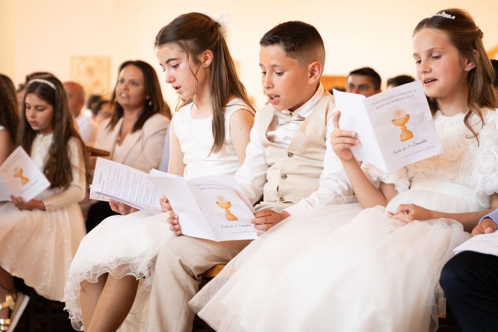 Children in white formal attire reading booklets at a First Communion ceremony in a decorated hall.