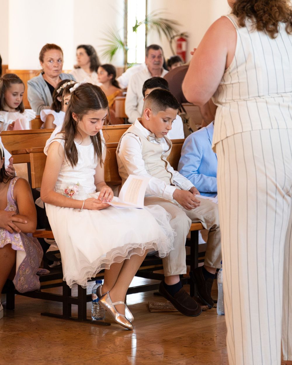 Children in white formal attire sit in church pews during a First Communion ceremony with family seated behind them.