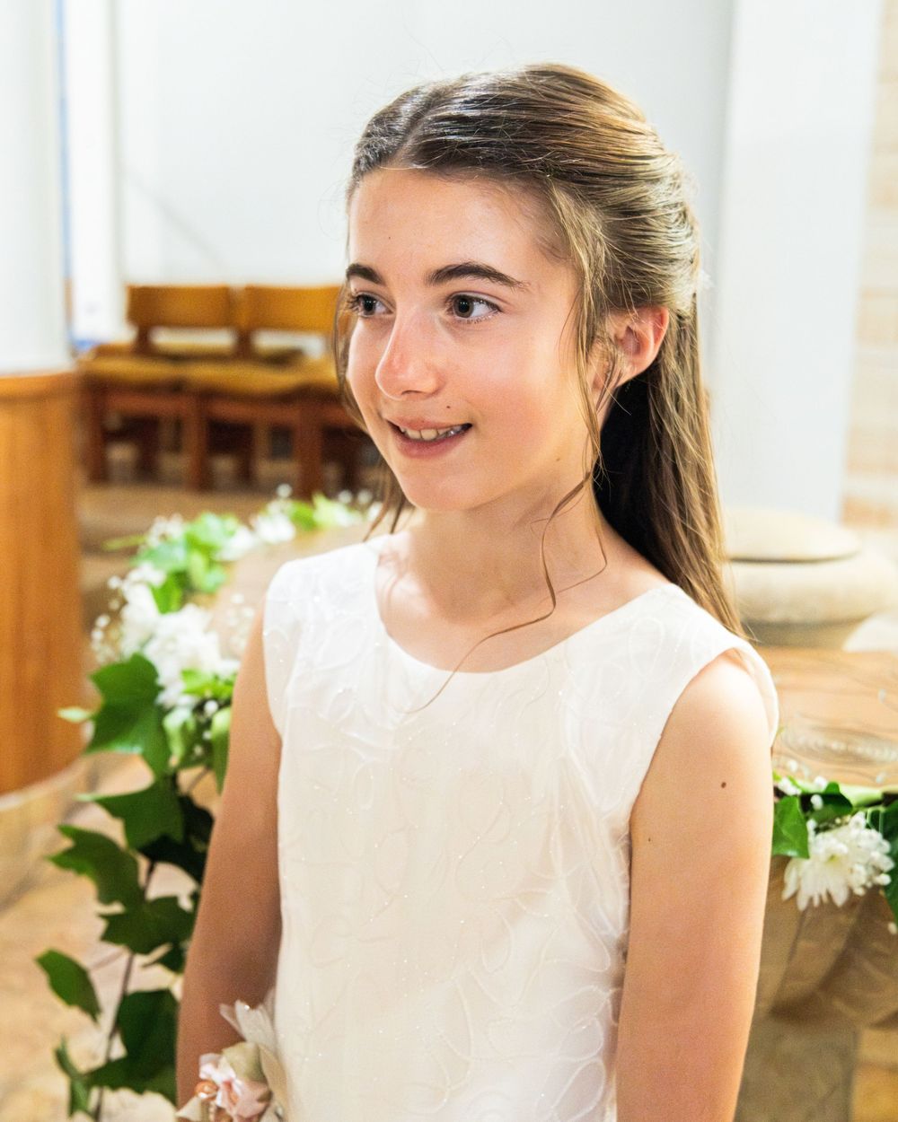 Young girl in white dress holding white flowers, smiling in a church setting during a special ceremony.