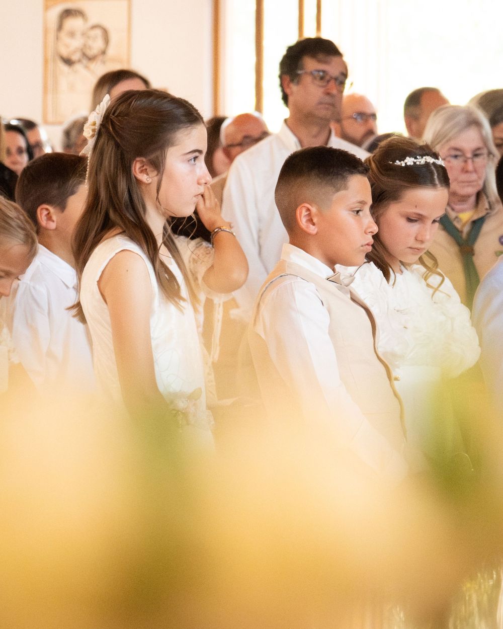 Children in white attire during a First Communion ceremony in a church, with adults gathered in the background.