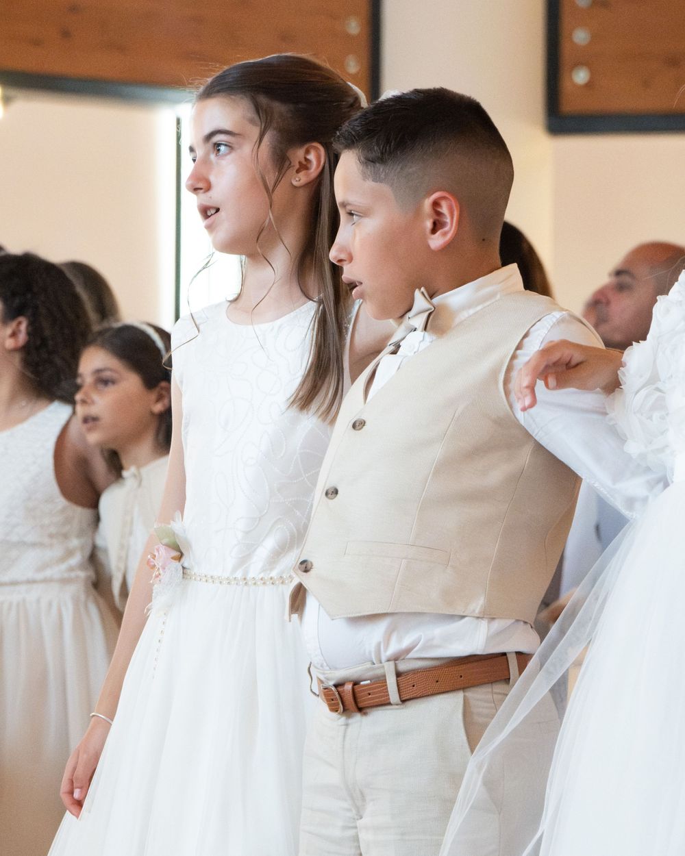 Children in white formal attire participate in a First Communion ceremony inside a church.