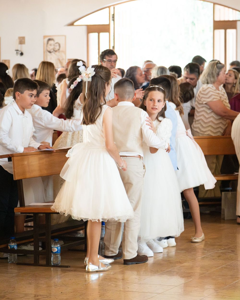 Children in white formal attire gather around church pews during a First Communion ceremony.