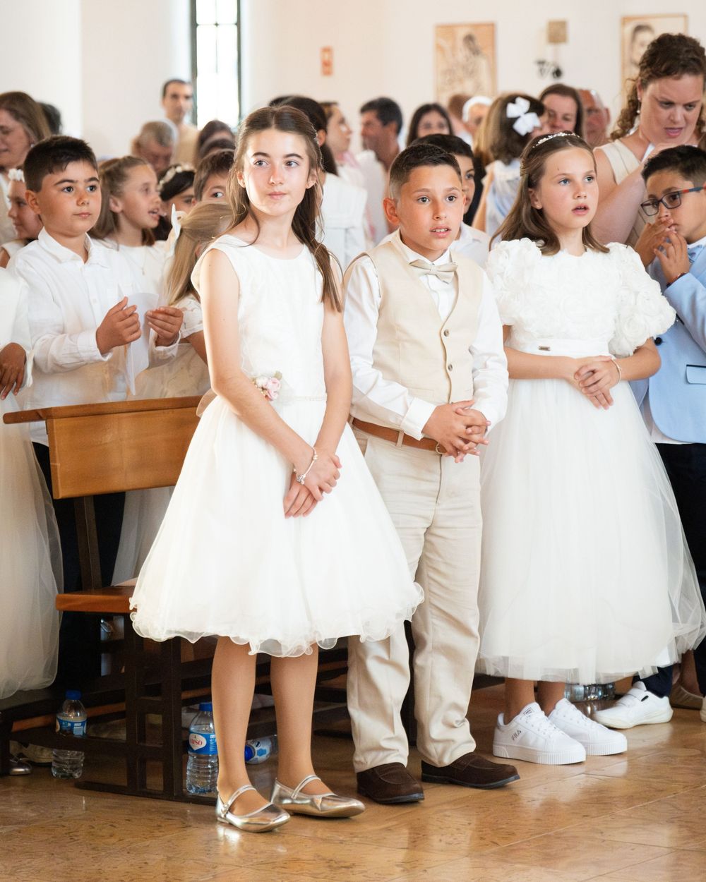 Children dressed in white formal attire stand together during a First Communion ceremony inside a church.