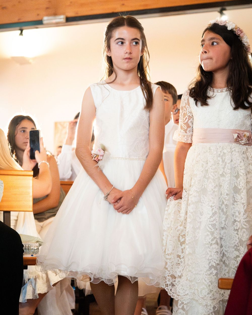 Two girls in white lace dresses stand during a First Communion ceremony inside a church.