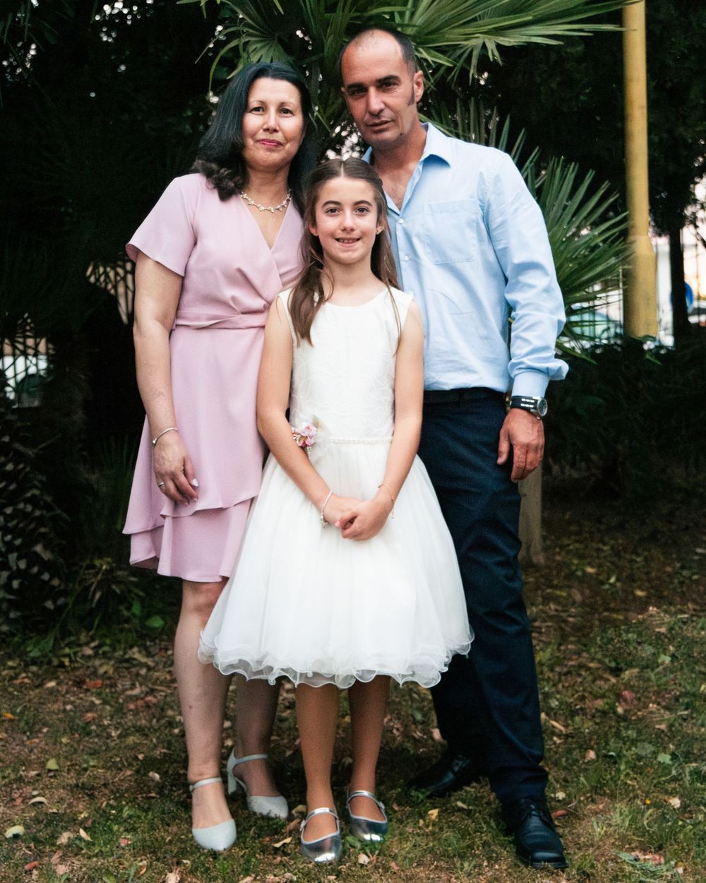 Family of three posing outdoors, woman in pink dress, girl in white tulle dress, man in blue shirt.
