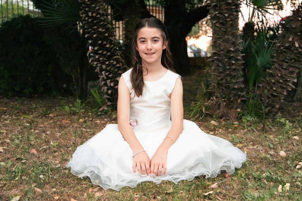 Young girl in white dress sitting on grass outdoors with palm trees and pine cones in background.