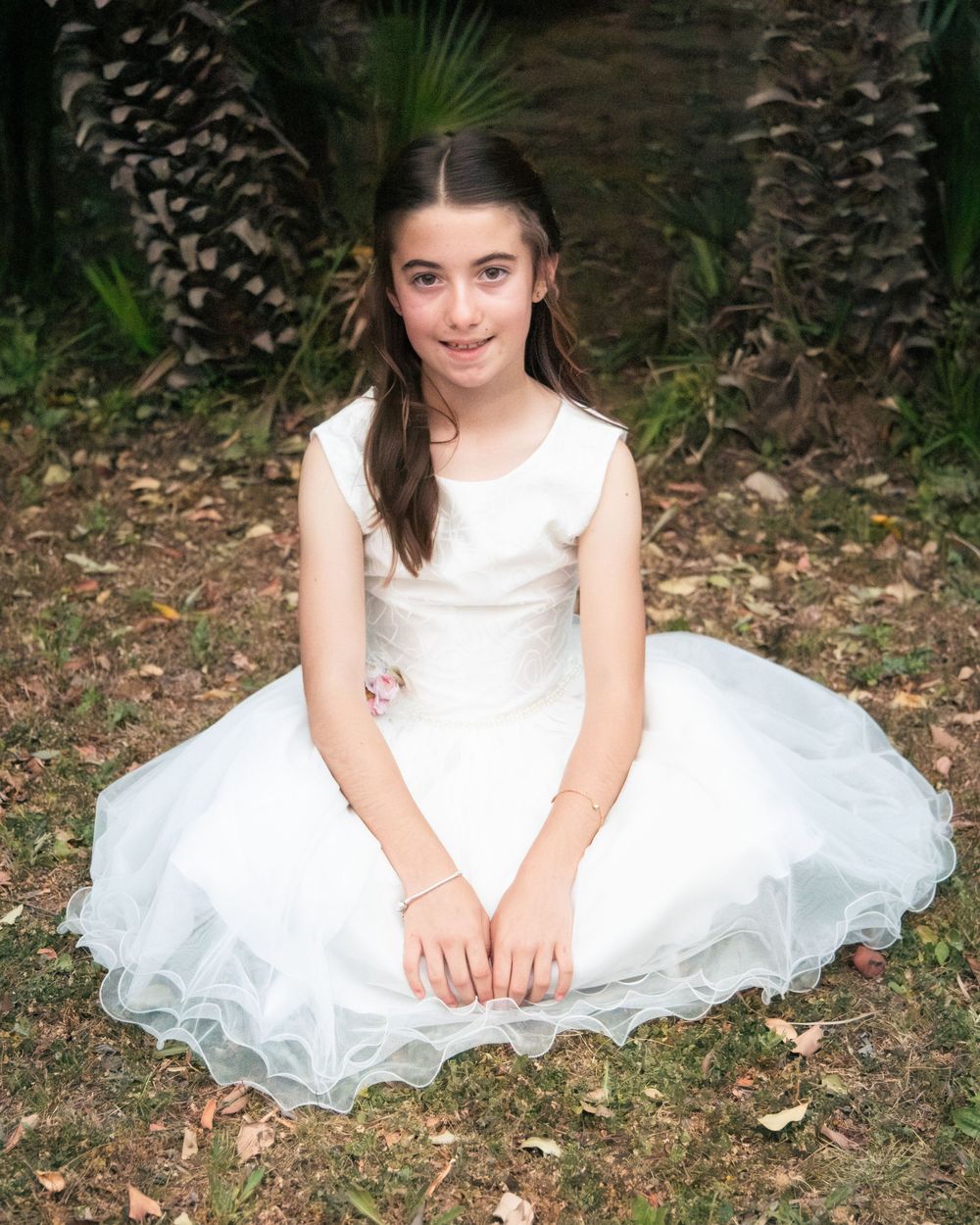 Young girl in white tulle dress sitting on grass outdoors, surrounded by palm trees, for a formal occasion.