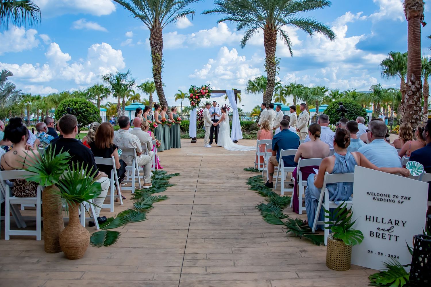 Outdoor wedding ceremony on wooden aisle lined with white chairs and palm trees at a tropical Florida venue.