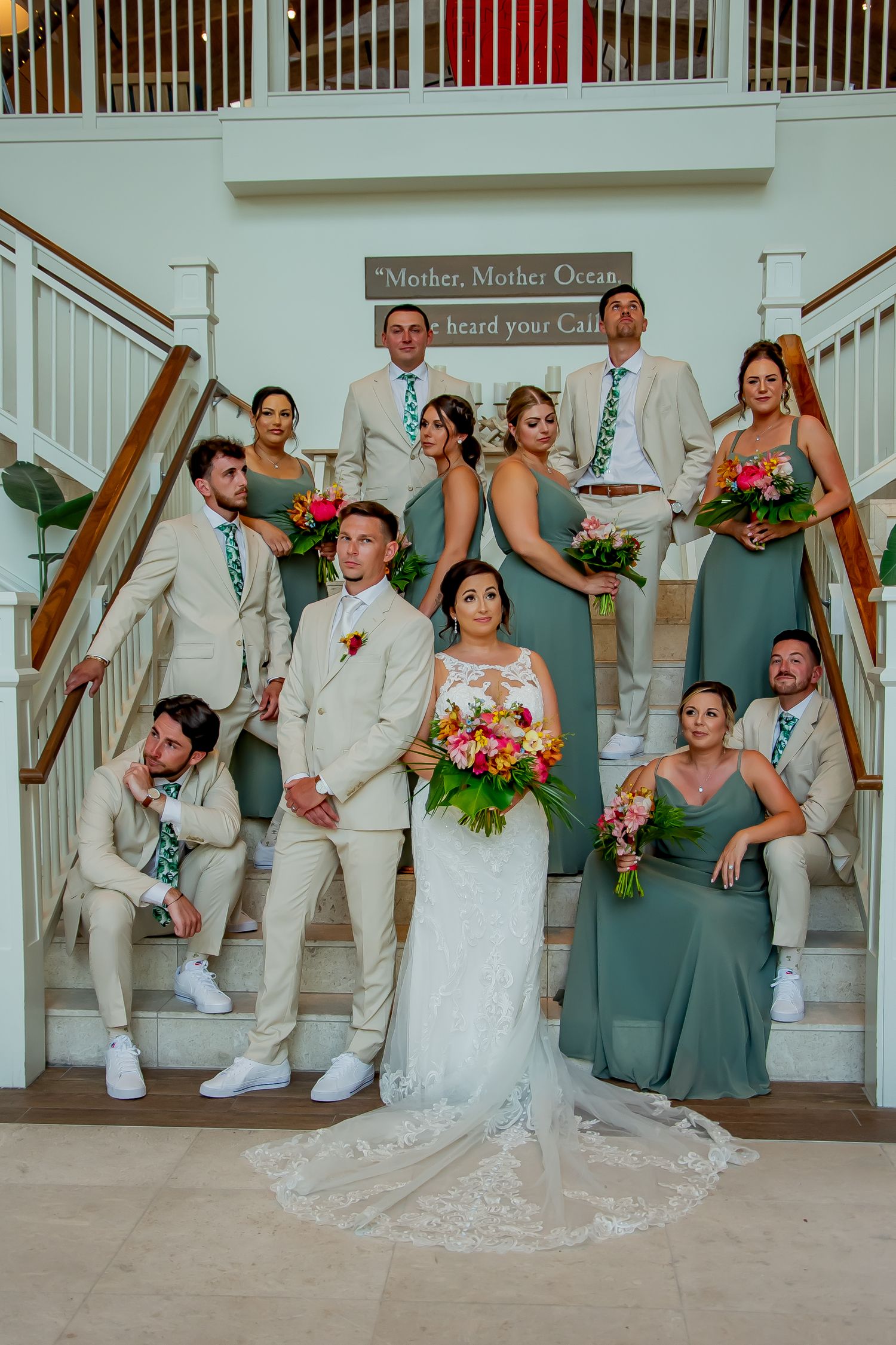 Wedding party poses on grand staircase, bride and groom centered, groomsmen in tan suits, bridesmaids in sage green dresses.