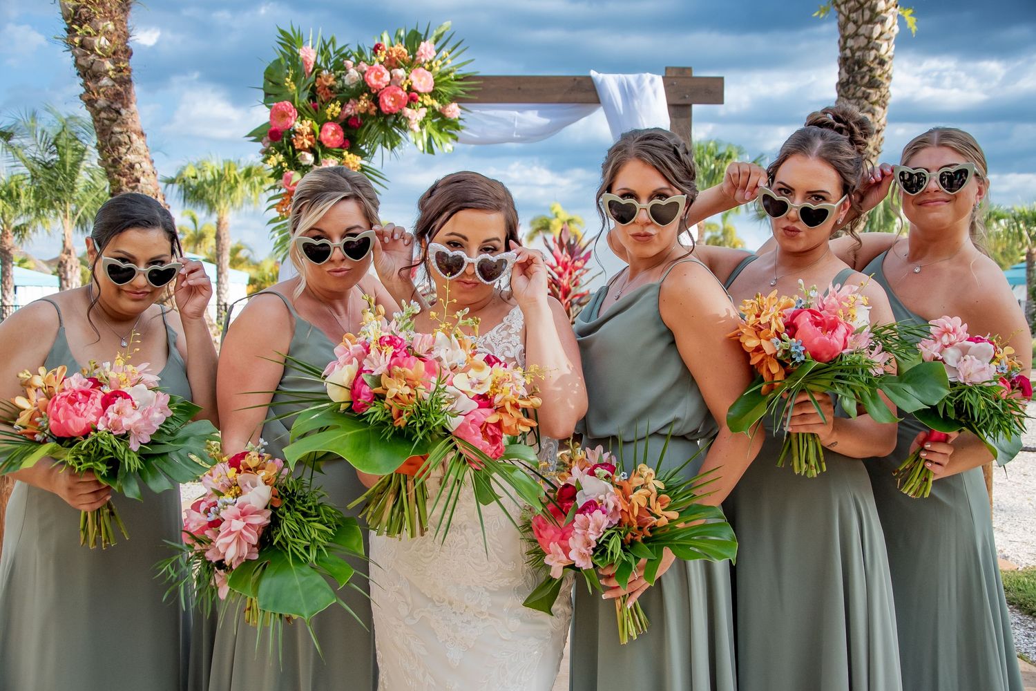 Bridesmaids in sage green dresses and white sunglasses hold tropical bouquets with bride at outdoor beach wedding.
