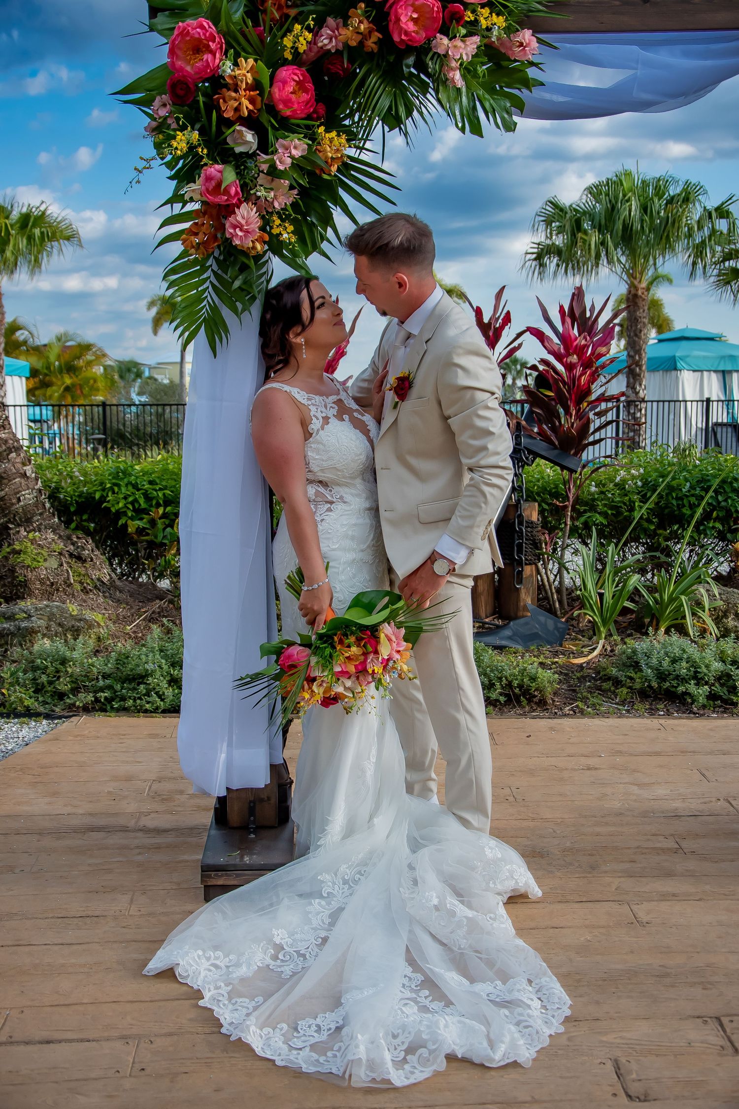 Bride and groom share a romantic moment under a floral arch with tropical flowers at an outdoor wedding ceremony.