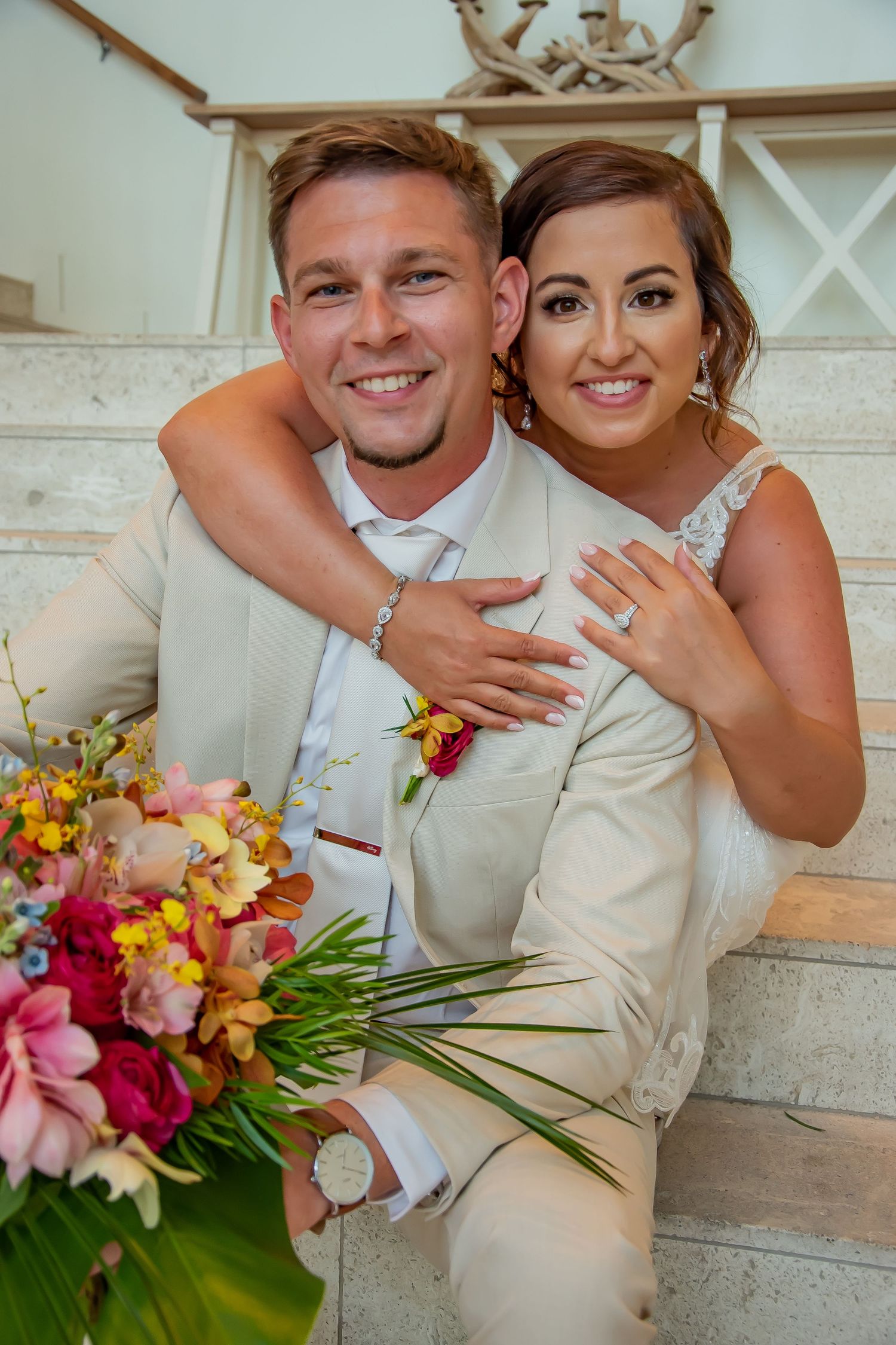 Smiling bride and groom in cream suit pose on marble steps with colorful tropical wedding bouquet.