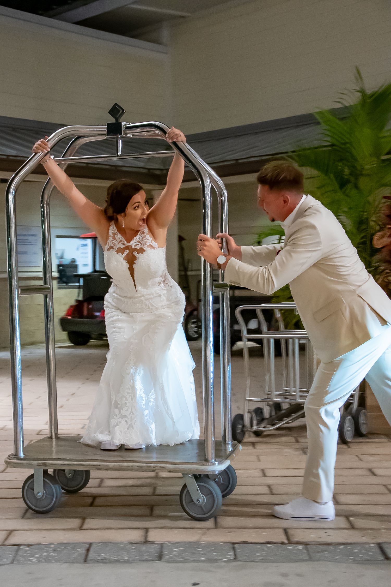 Bride in white gown rides hotel luggage cart pushed by groom in cream suit during fun wedding photo.