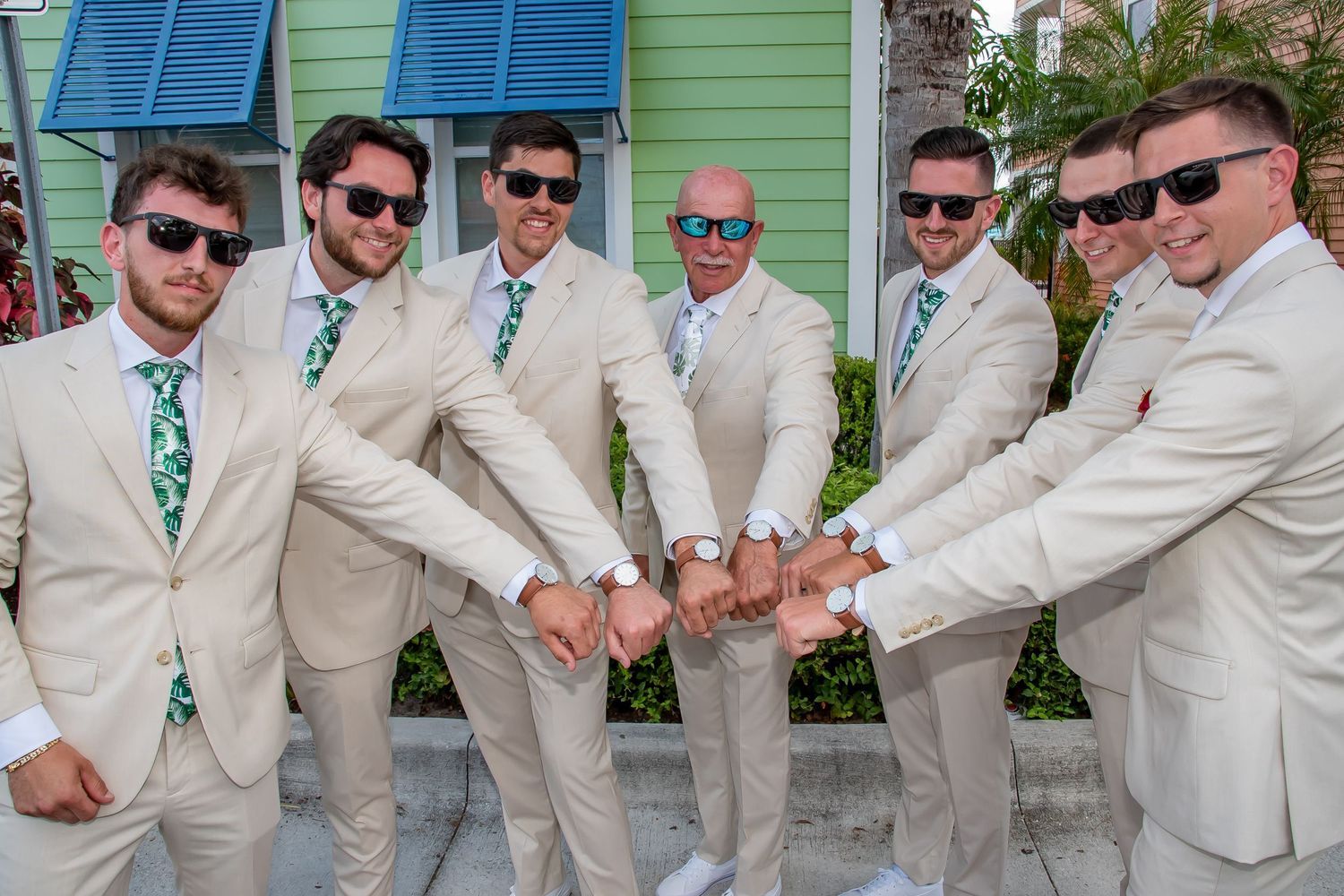 Six groomsmen in matching tan suits and green ties show off their watches outdoors at a sunny wedding.