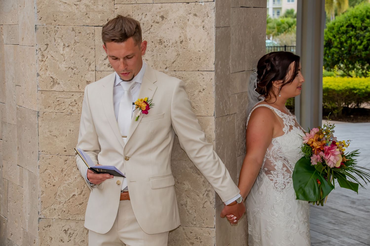 A groom in a white suit reads vows while holding his bride's hand before their outdoor wedding ceremony.