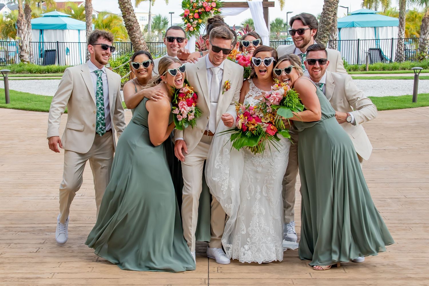 Wedding party in sage green and tan attire posing together outdoors, bride holding tropical bouquet, all wearing fun sunglasses.