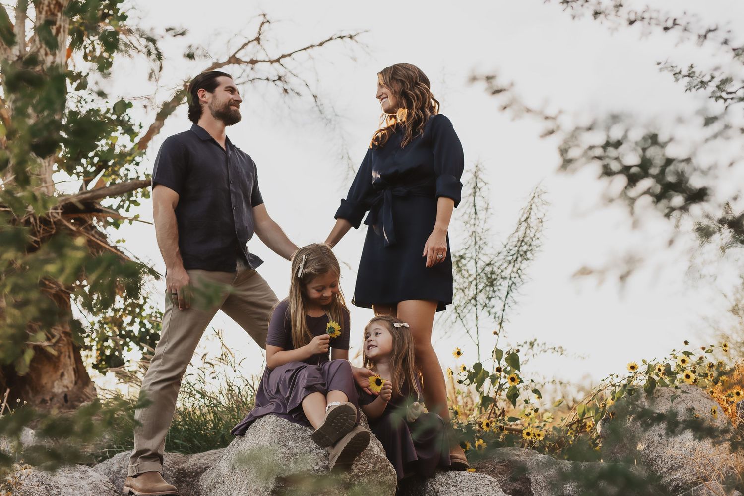 Family of four posing outdoors on rocks surrounded by trees, parents standing while two children sit below them.