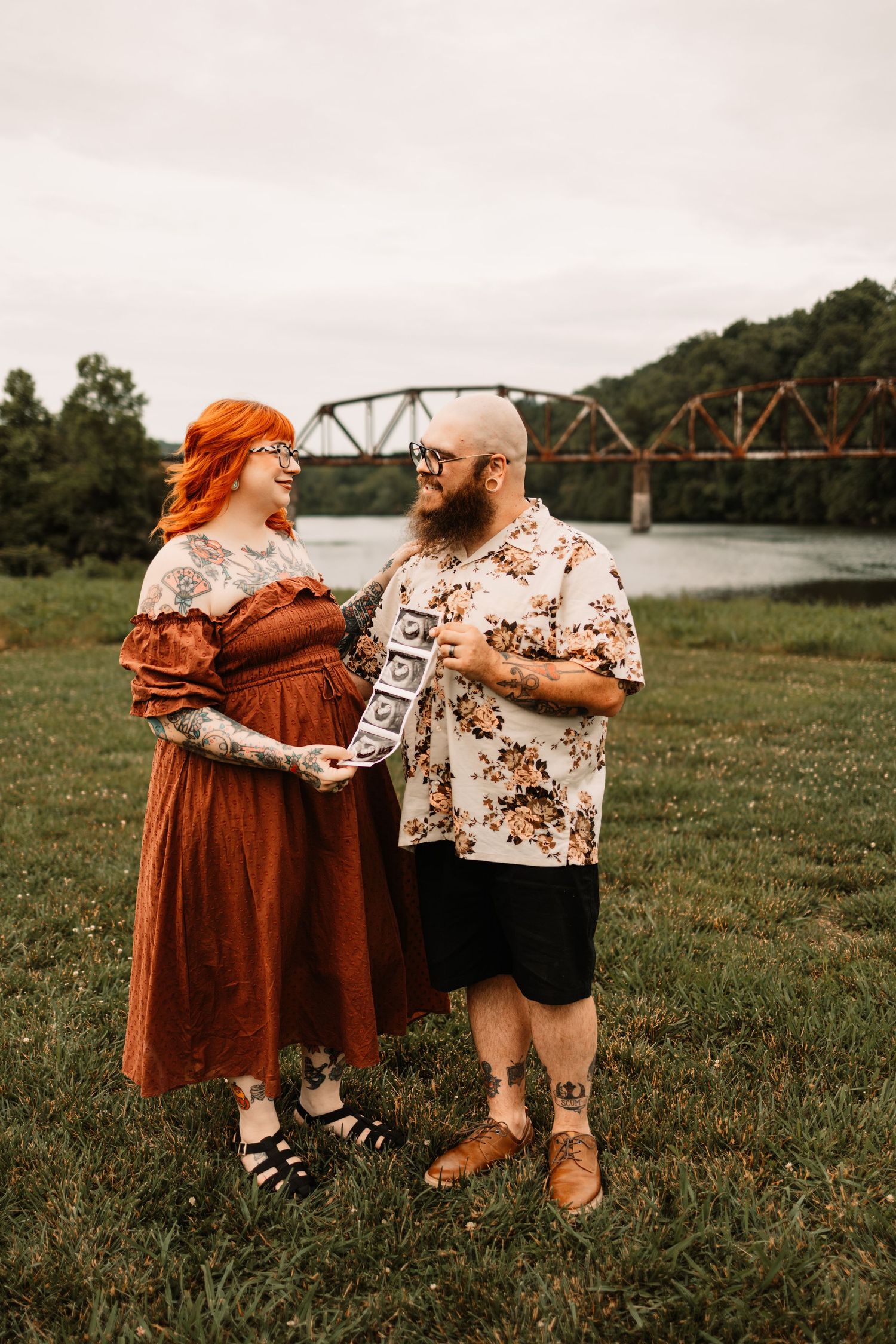 Couple holding ultrasound photos outdoors near a river bridge, woman in rust dress with red hair, man in floral shirt.