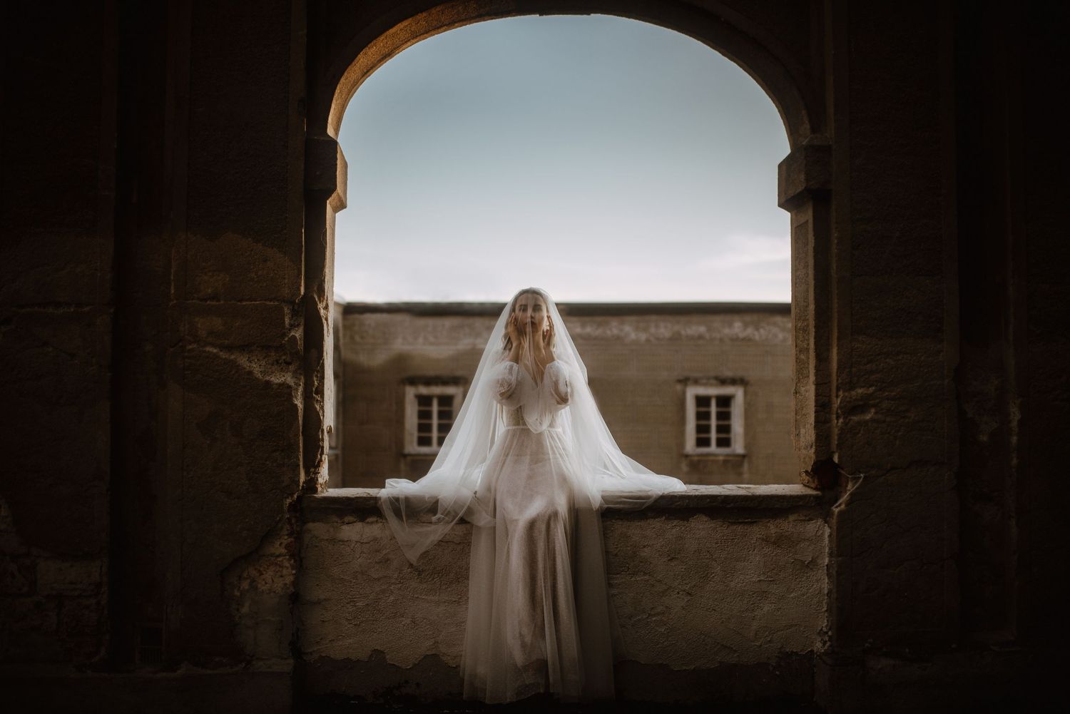 Bride in flowing white gown and veil stands in stone archway, backlit by soft sky at historic venue.