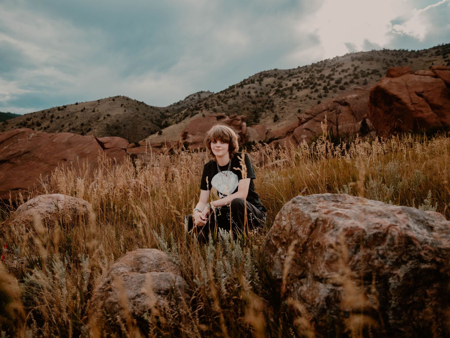 Boy crouches in grass and poses for senior photos, cinematic, mountain, and portrait photos in Morrison, Colorado outside and in nature