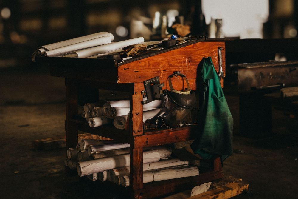 Rustic wooden workbench with tools, rolled blueprints, and a green cloth in a dimly lit workshop.