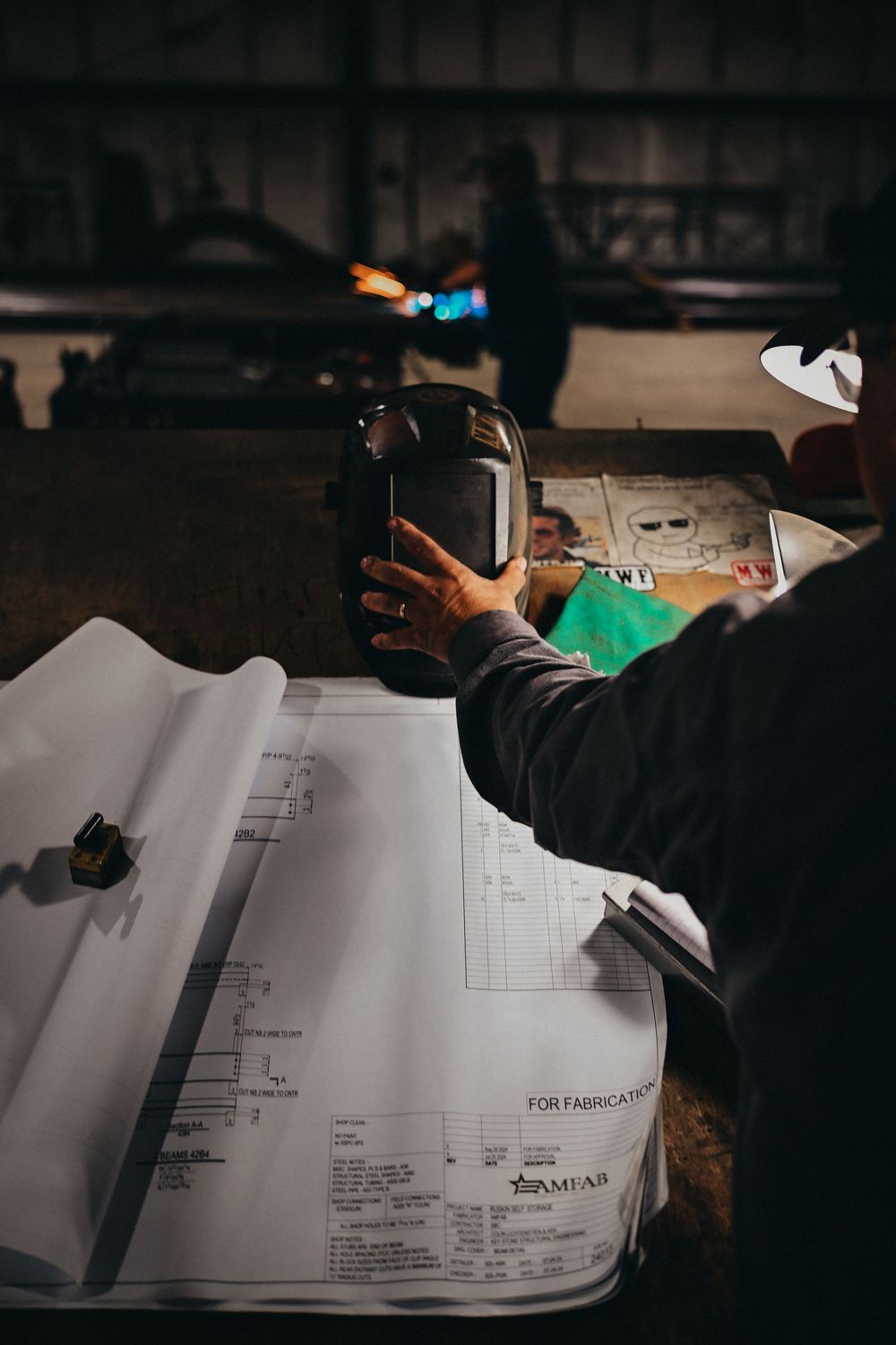 Welder holding a welding helmet over fabrication blueprints in an industrial workshop.