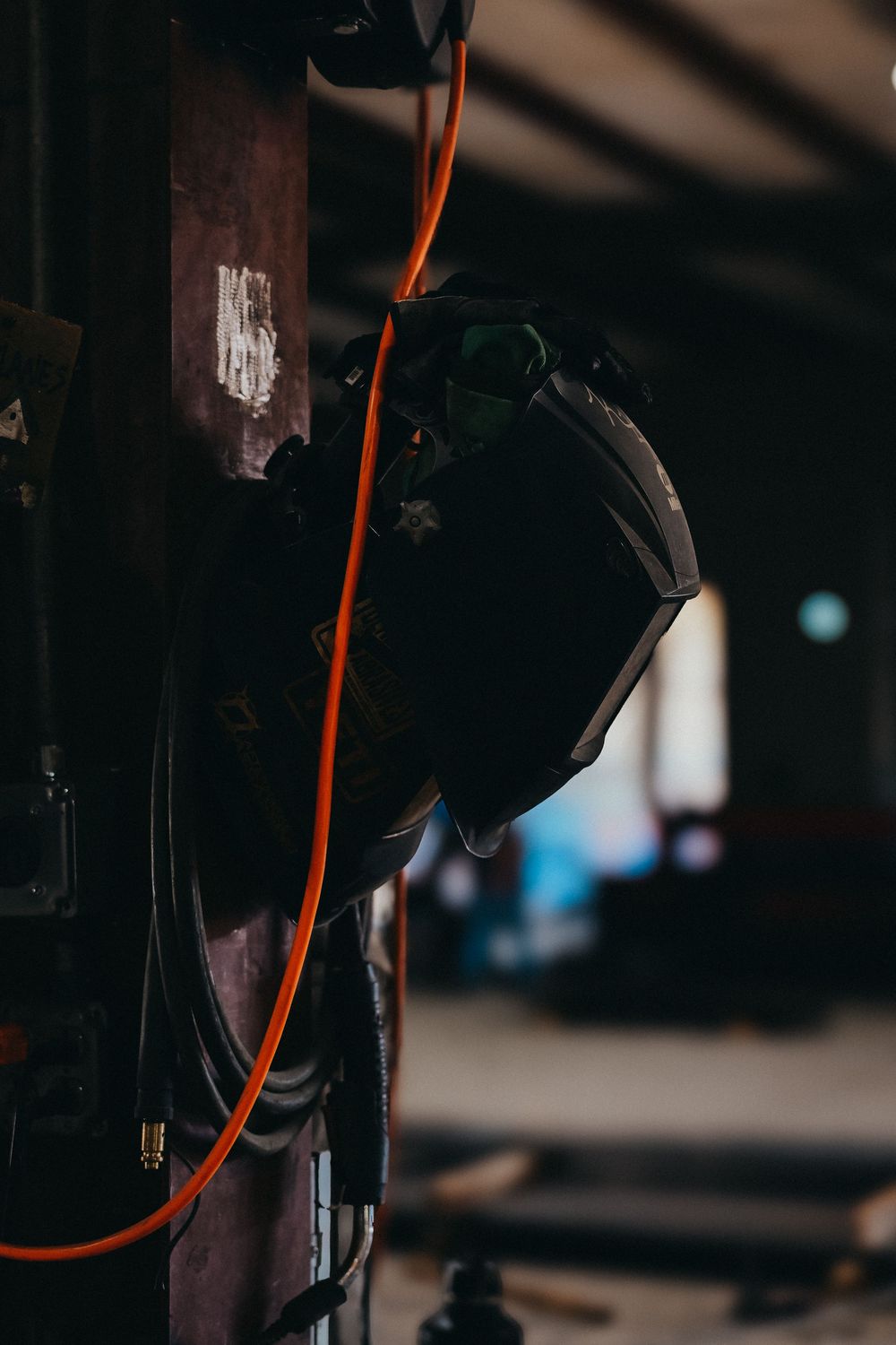 A welding helmet hanging on a dark metal post with an orange air hose in an industrial workshop setting.