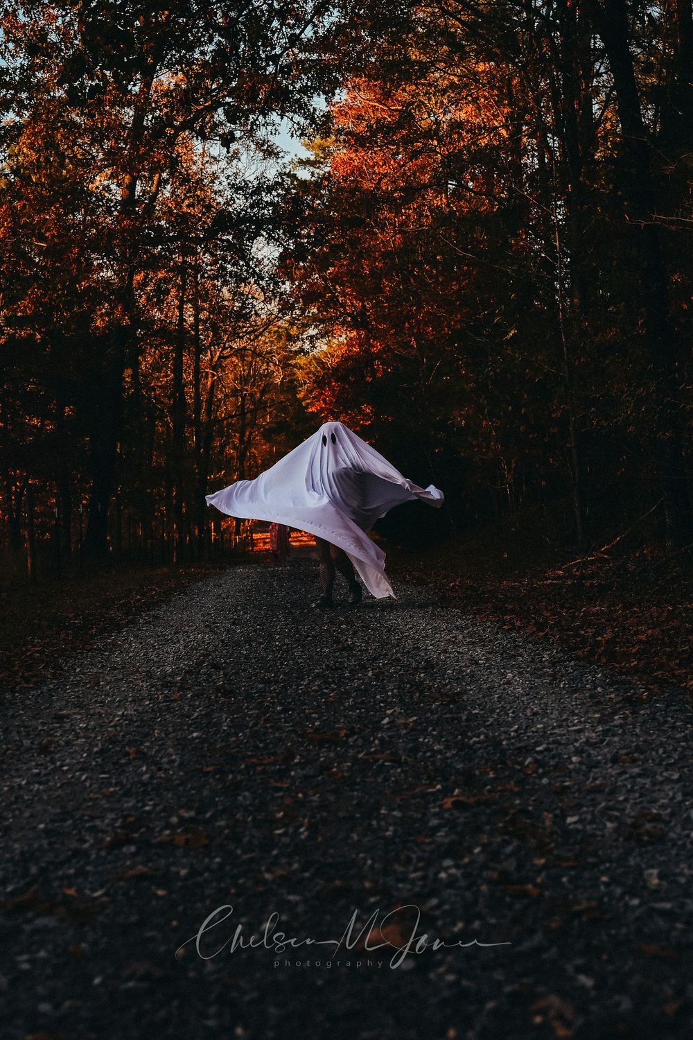 Person dressed as a ghost on a gravel path surrounded by vibrant autumn foliage in a dark forest setting.