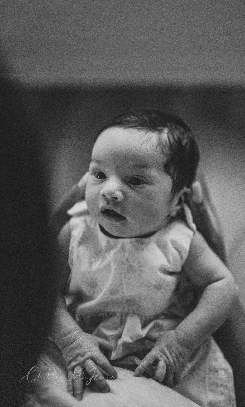Black and white photo of a newborn baby with dark hair being held by an adult, wearing a floral onesie.