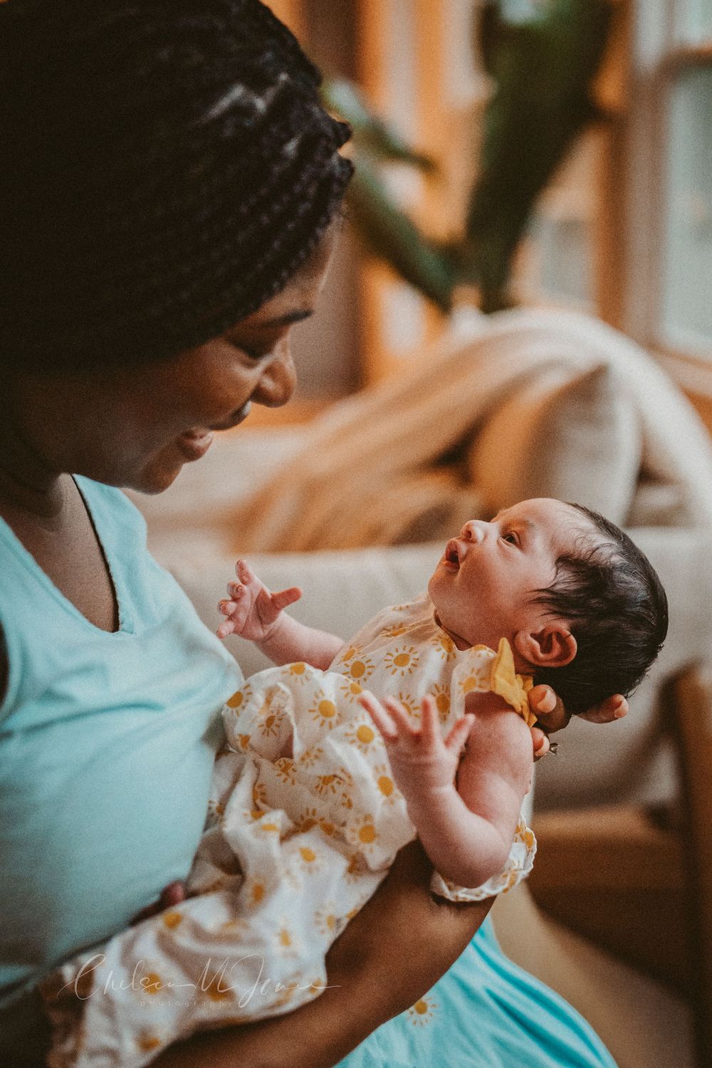 A Black mother tenderly holds her newborn baby in a sunlit room, both dressed in light blue and white floral outfits.
