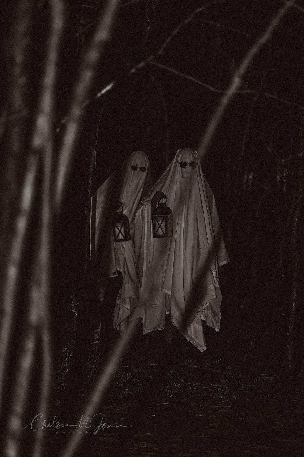 Two ghost costumes with glowing eyes carry lanterns through a dark spooky forest in a black and white Halloween photo.